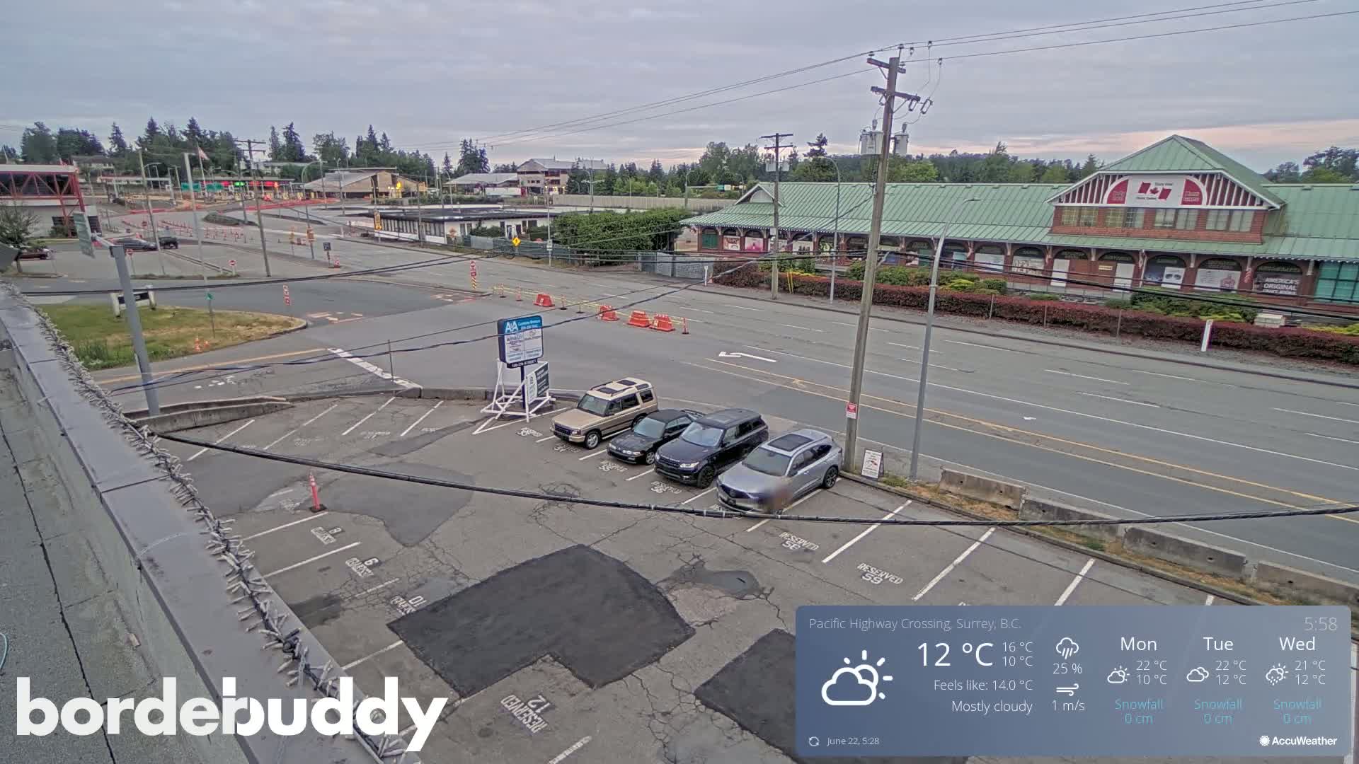 A mostly cloudy daytime scene shows a parking lot with several cars, a road with light traffic, and buildings in the background.