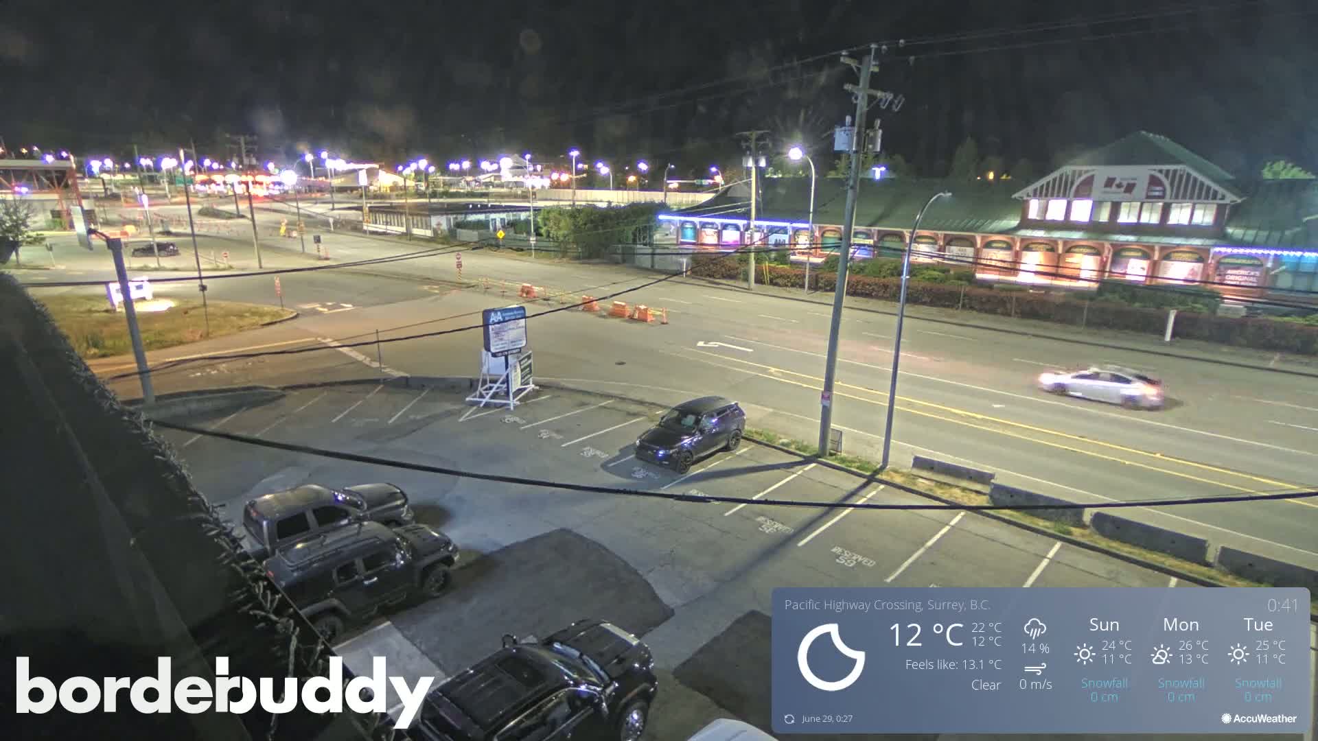 A nighttime view of a highway crossing, parking lot, and building with several cars visible under clear skies.