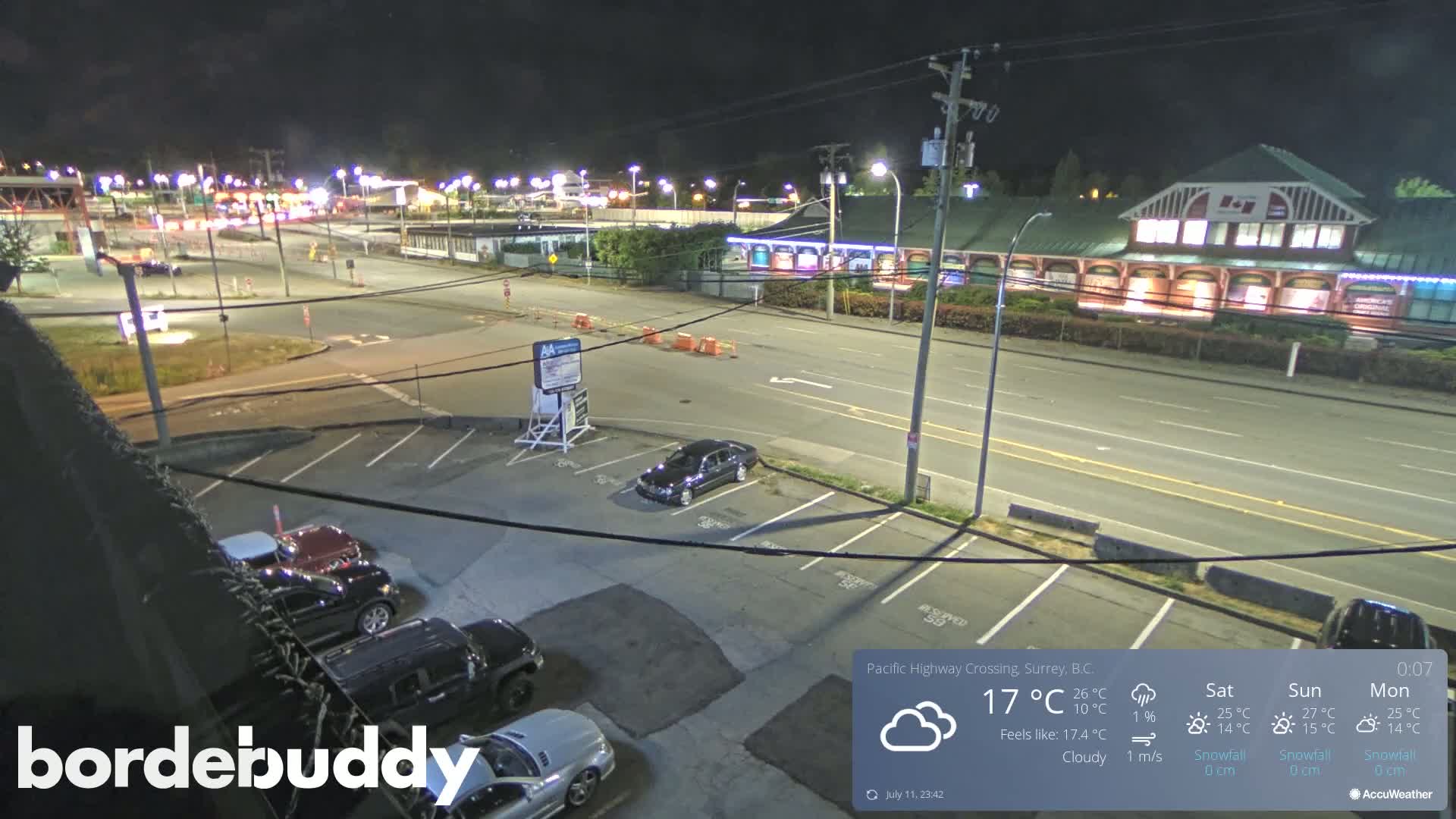 A nighttime, cloudy view of a parking lot and roadway with several cars parked, leading to a brightly lit building across the street.