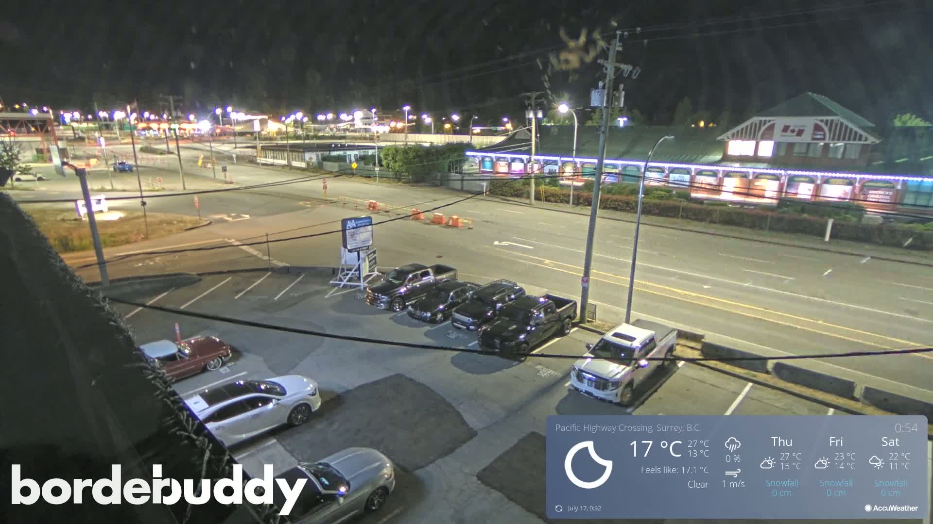 A nighttime view of a parking lot and roadway with several cars parked, under clear skies.