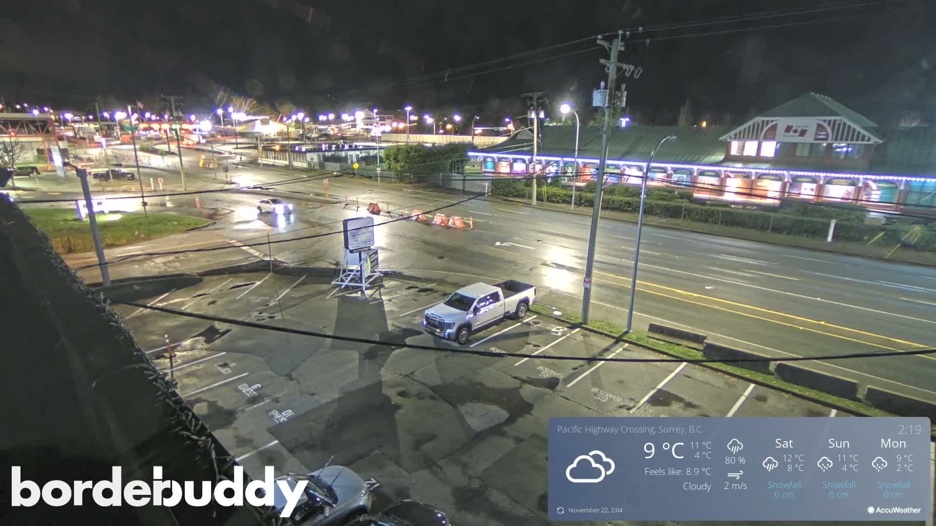 An urban intersection and parking lot are seen at night under damp conditions, with a distinctive brightly lit building on the right and a white pickup truck parked in the foreground, all reflecting artificial lights on the wet pavement.