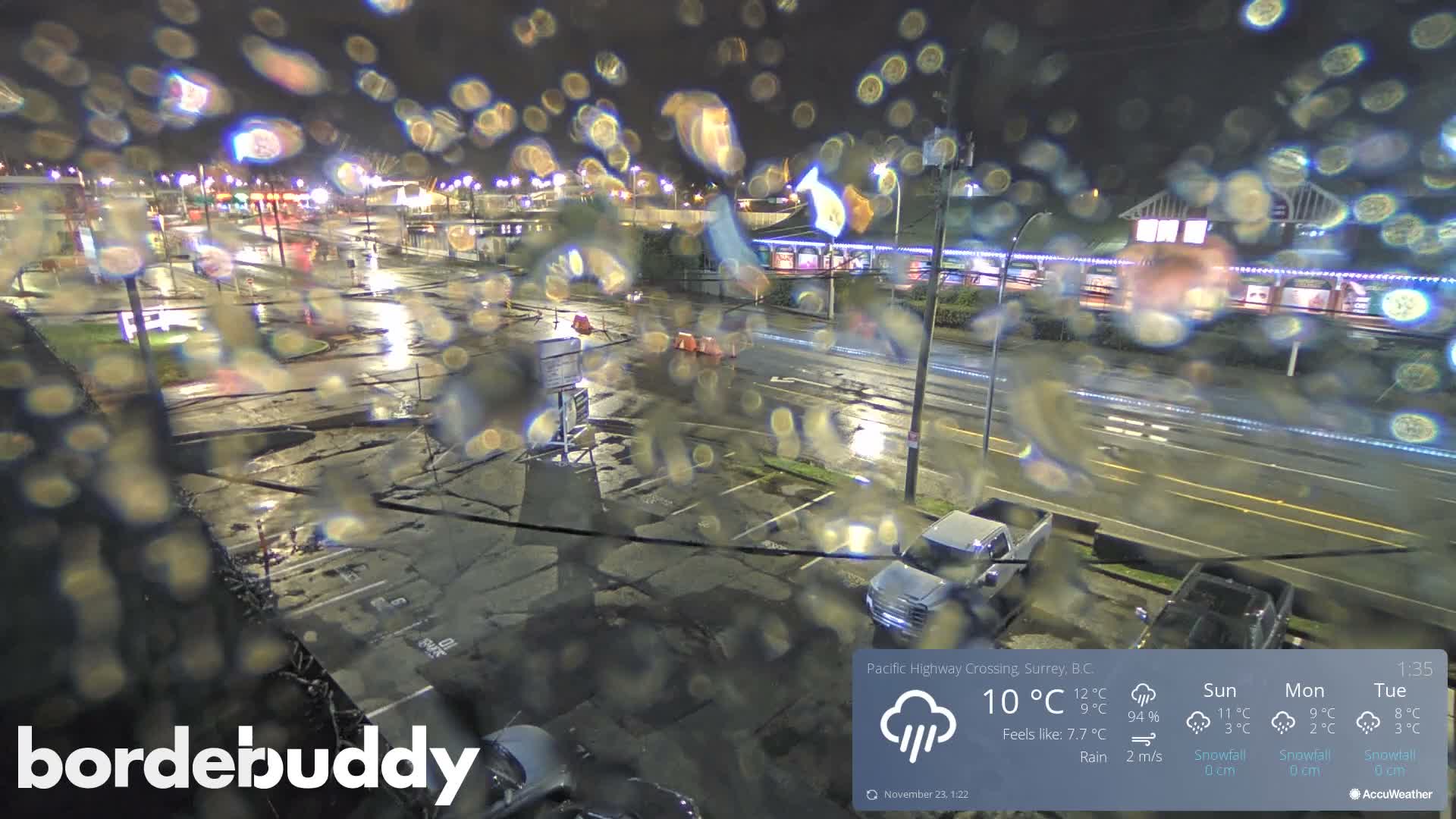 The image shows a rain-soaked outdoor area at night with wet pavement reflecting lights from distant commercial buildings and parked vehicles, all viewed through a heavily water-splattered window.