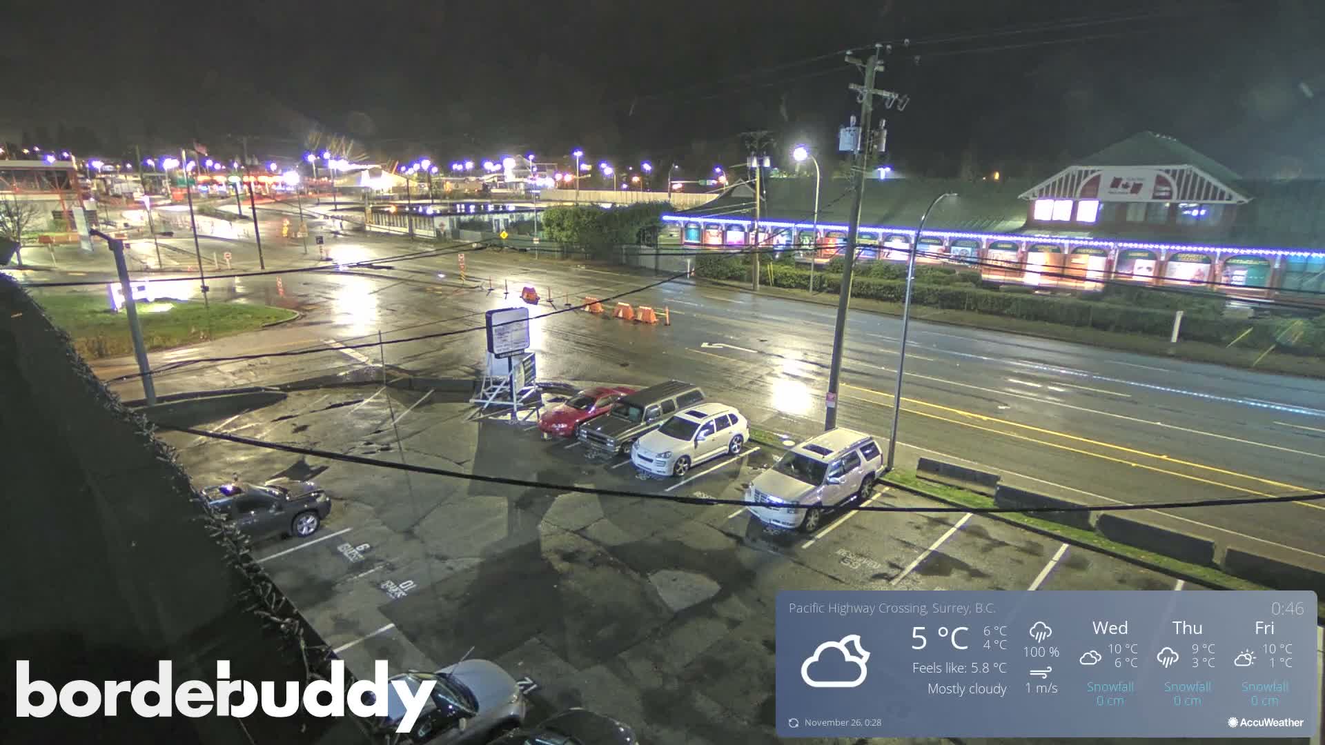 A nighttime aerial view captures a wet parking lot with cars and a multi-lane road lined by illuminated commercial buildings, reflecting the streetlights on the rain-slicked surfaces.