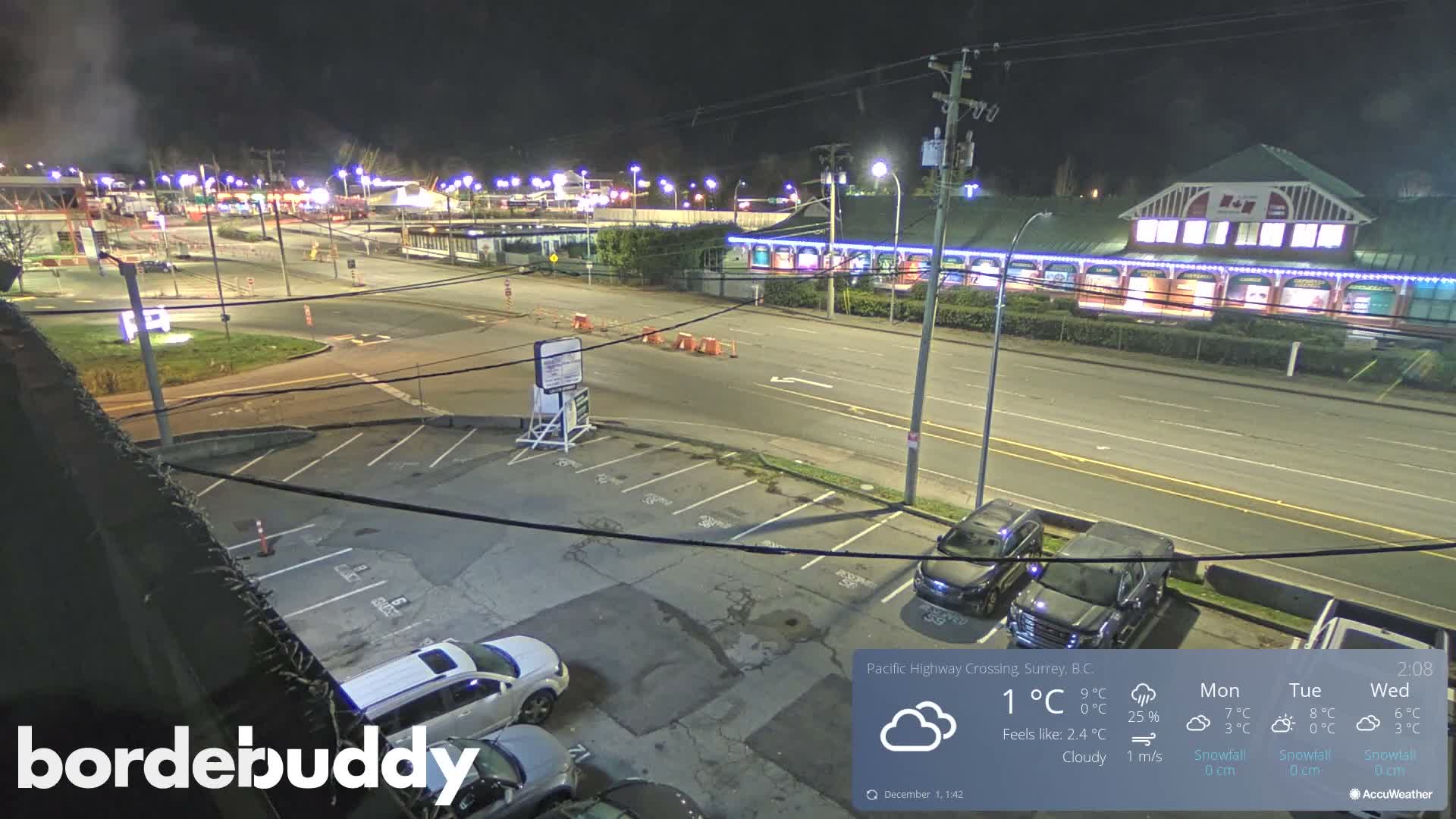 The image displays an elevated nighttime view of a multi-lane road bordered by a brightly lit commercial building and other establishments, with a parking lot containing several cars in the foreground under cloudy skies.