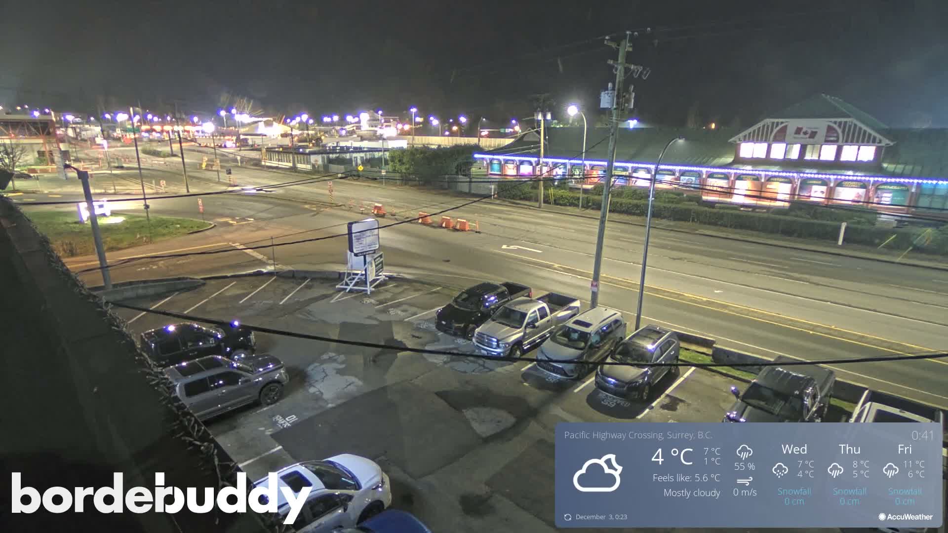 A night-time view shows a well-lit commercial area with a parking lot full of cars in the foreground, a multi-lane road, and brightly illuminated buildings featuring blue decorative lights in the background, all under a dark sky with visibly wet ground conditions.