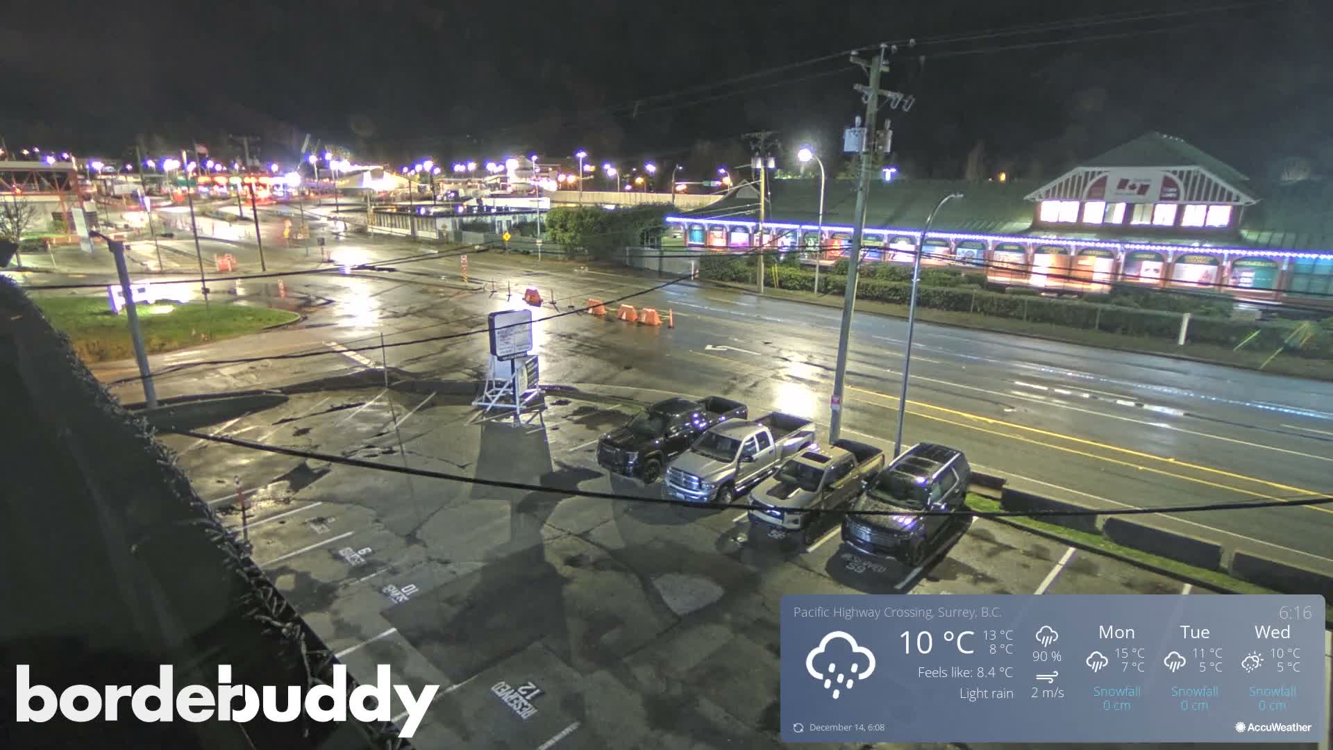 A night-time view shows a well-lit commercial area with a parking lot full of cars in the foreground, a multi-lane road, and brightly illuminated buildings featuring blue decorative lights in the background, all under a dark sky with visibly wet ground conditions.