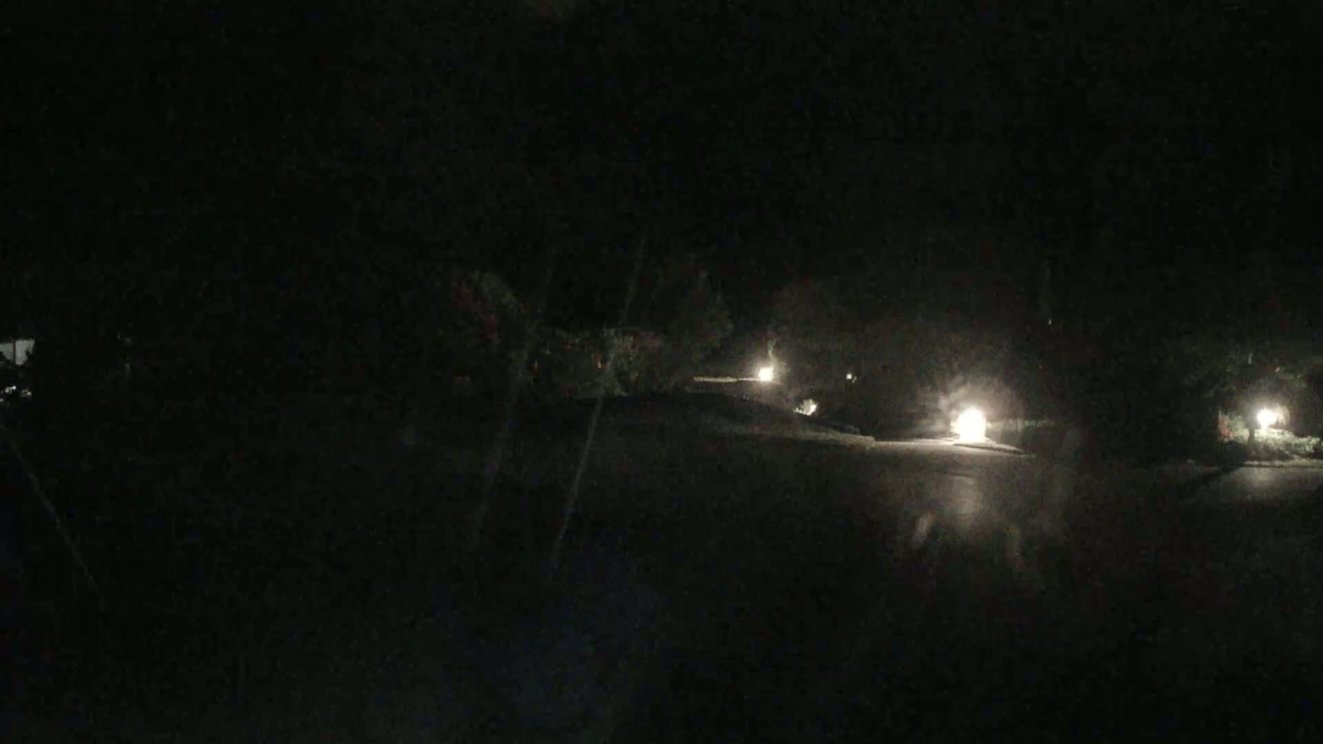 A dark, nighttime scene shows a paved road dimly lit by several small lights along the roadside, surrounded by dark vegetation.