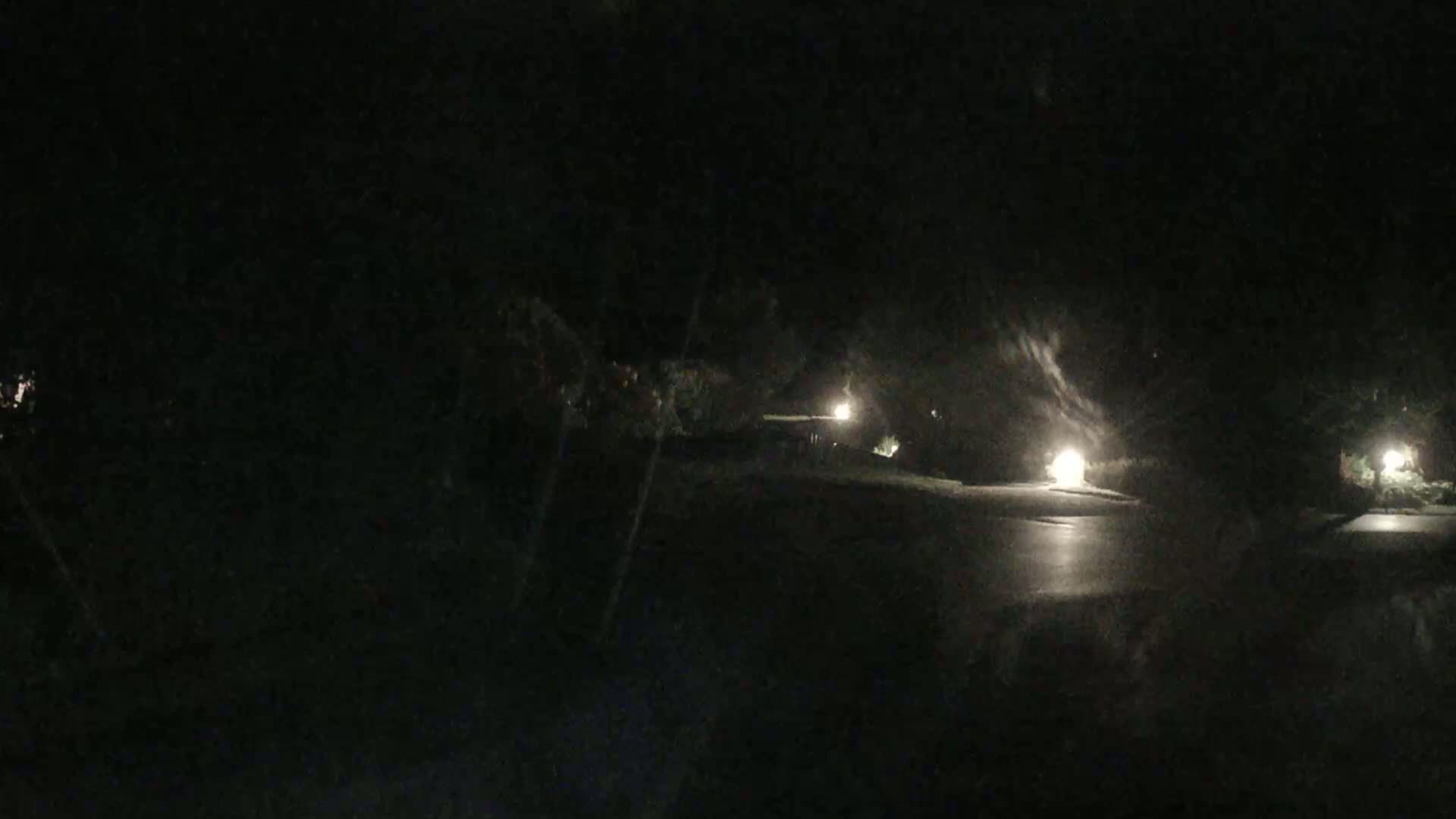 A dark, nighttime scene shows a wet, paved area illuminated by several small lights surrounded by dark trees.