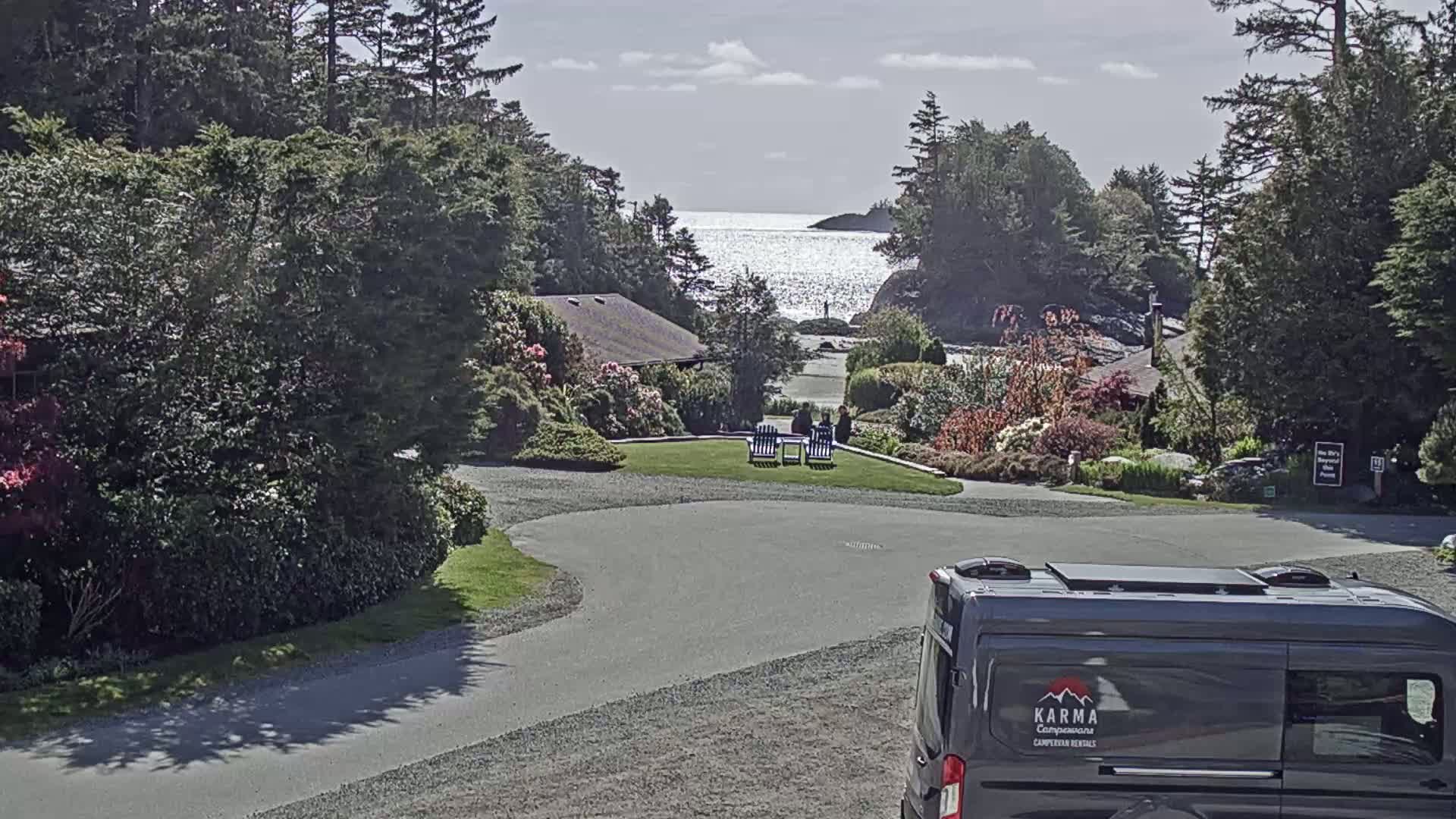 A sunny day reveals a coastal scene with a calm ocean view, a grassy area with seating, lush green landscaping, and a large gray campervan parked nearby.