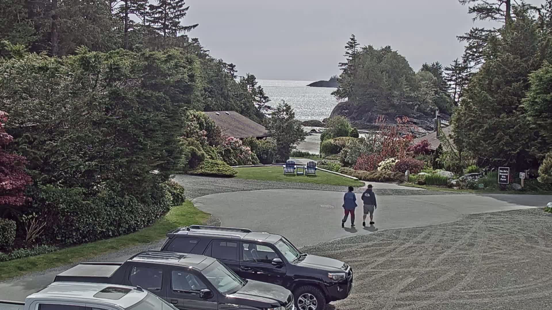 A gravel parking lot with two people walking, several parked vehicles, lush greenery, a small building, and a calm ocean view in the background under a mostly cloudy sky.