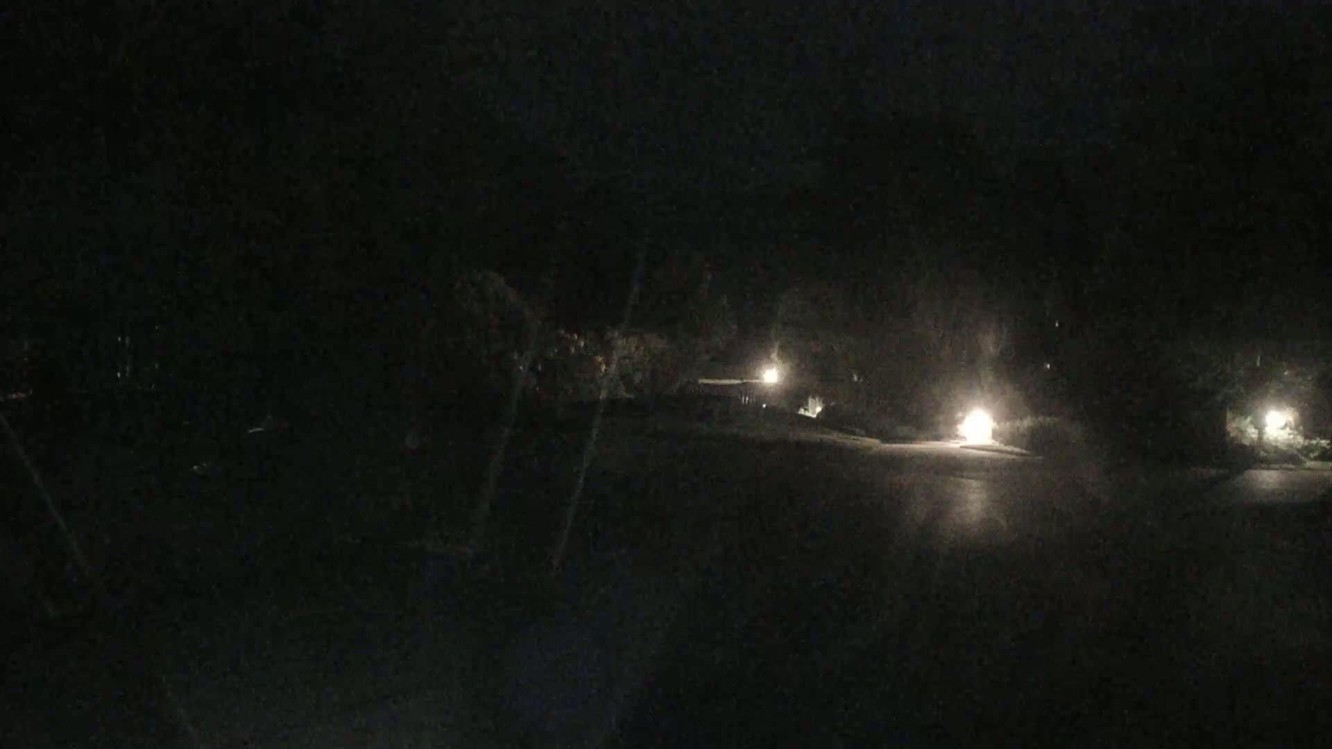 A dark, nighttime scene shows a paved road with sparsely lit landscaping and the headlights of a vehicle in the distance.