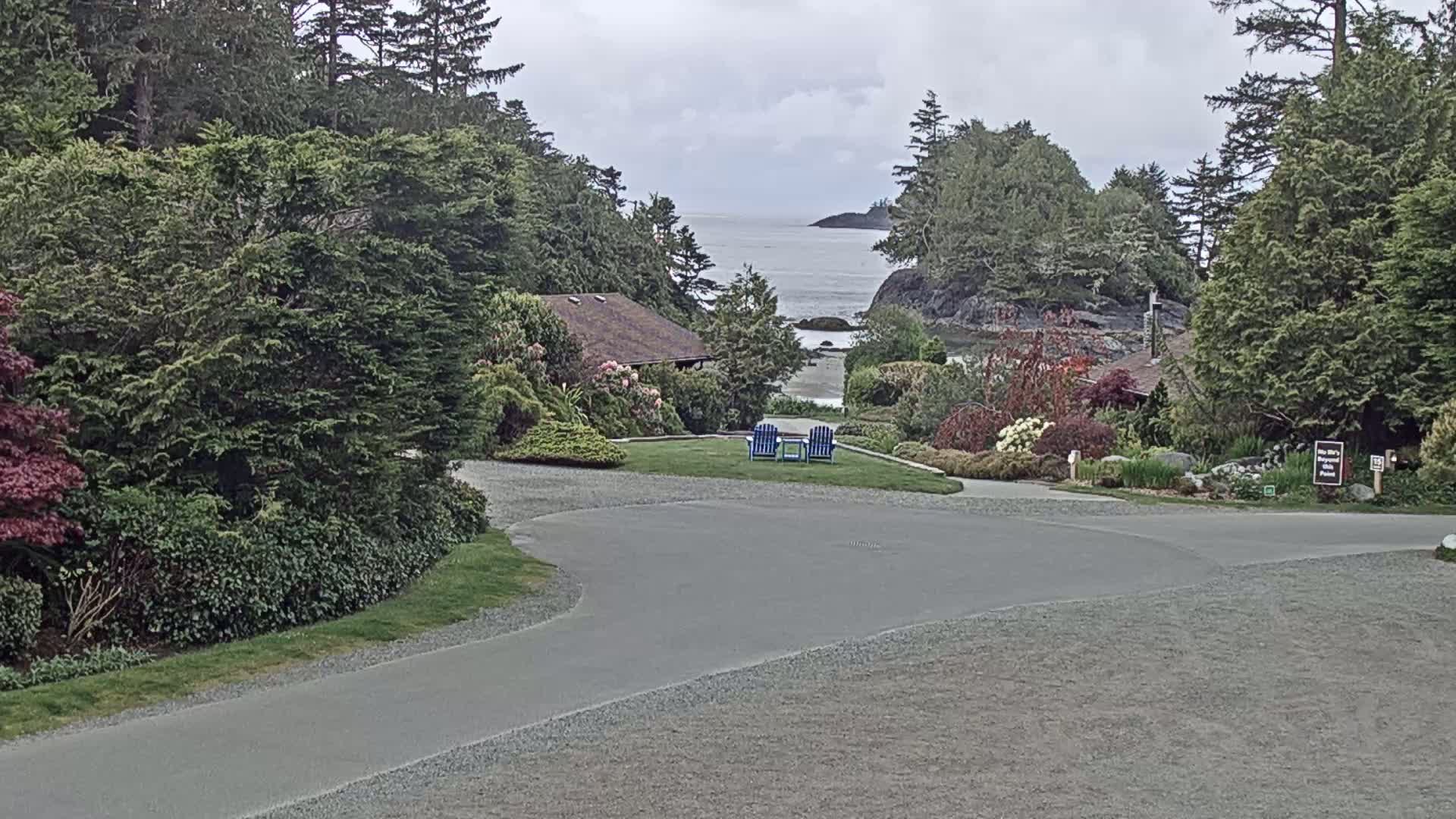A paved pathway curves through a lush, green landscape featuring a house, manicured gardens, and two blue chairs overlooking a calm ocean under a cloudy sky.