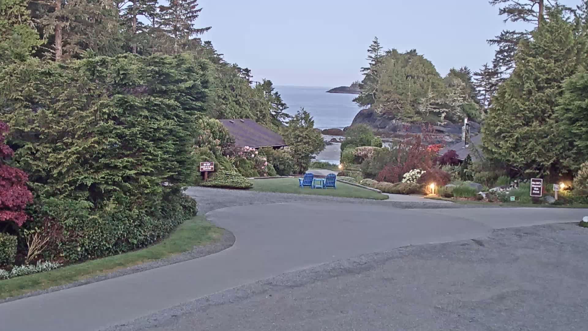 A paved pathway curves through a landscaped area with lush greenery, leading to a small building and a view of a calm ocean under a twilight sky.