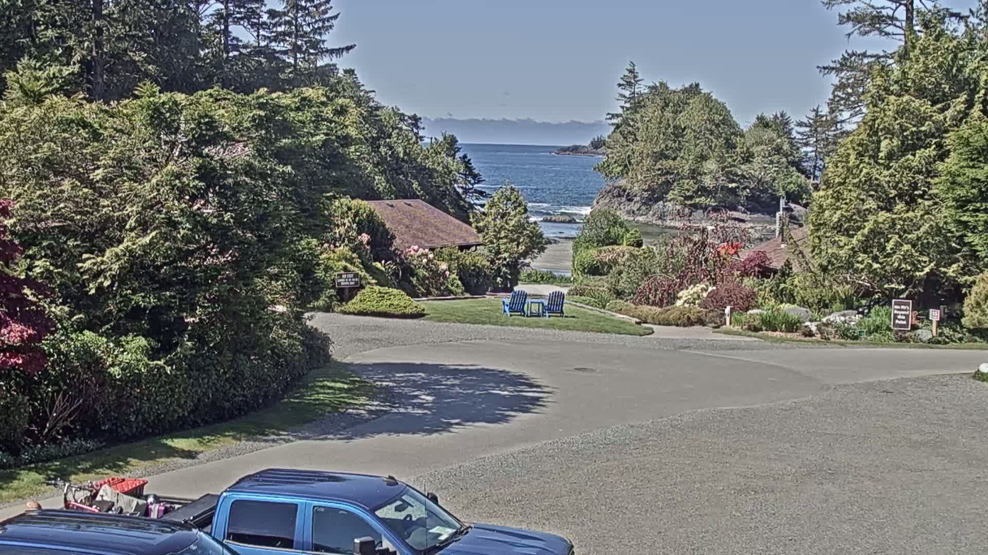 A paved parking lot overlooks a grassy area with two blue chairs, a small building, lush green landscaping, and a calm ocean with mountains in the distance under a sunny sky.