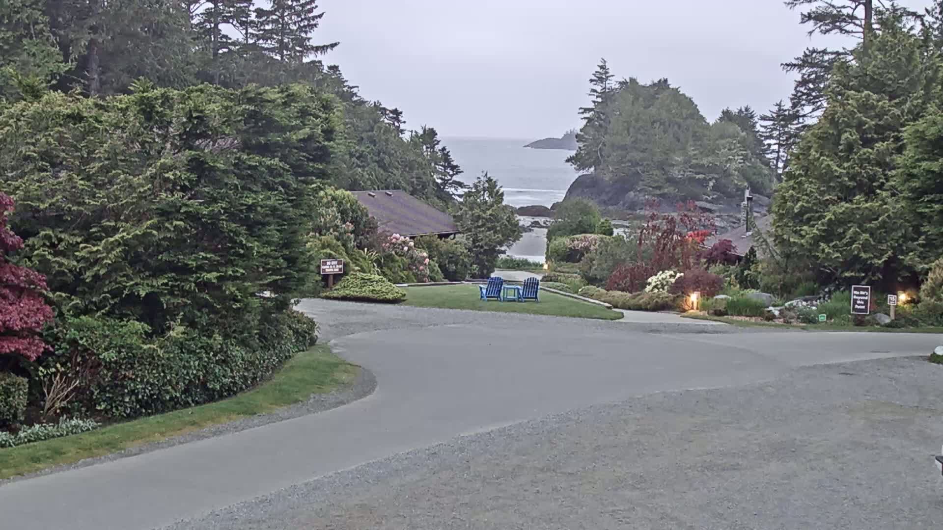 A paved driveway curves through a landscaped area with lush greenery, leading to a small building and a view of a calm ocean under an overcast sky.