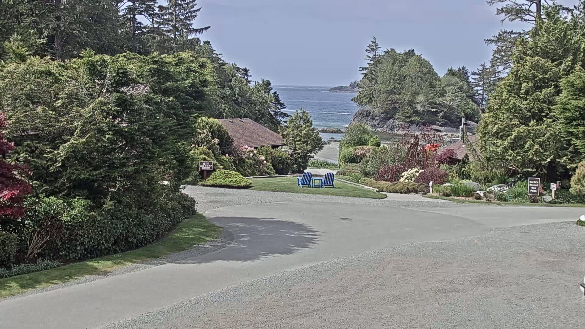 A paved roadway curves through a lush, green landscape featuring a small building, manicured lawns, and colorful flowerbeds, all overlooking a calm ocean on a partly sunny day.