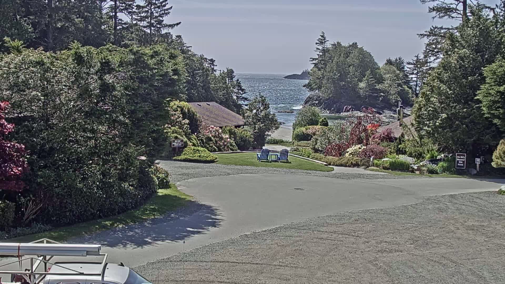 A paved area curves toward a grassy lawn with two blue chairs, overlooking a calm ocean and lush green vegetation under a sunny sky.