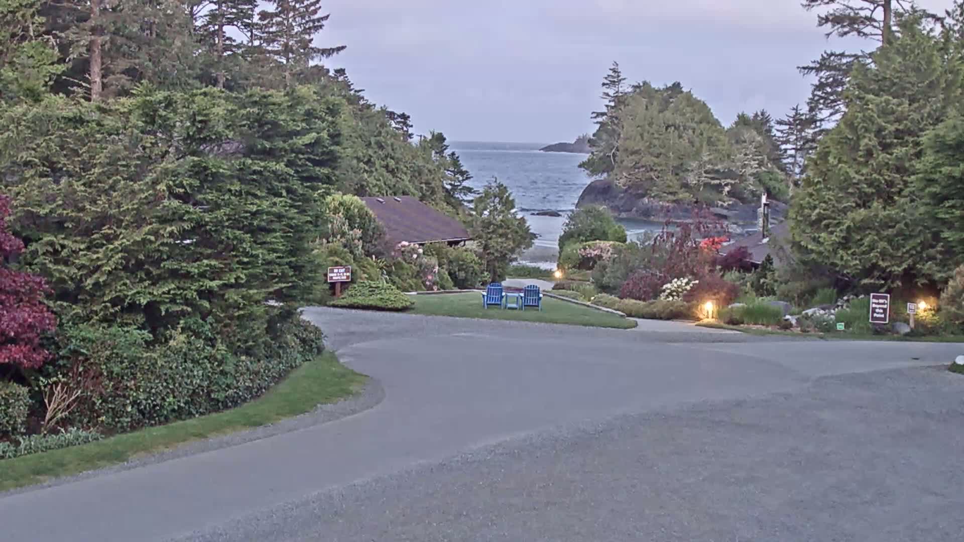 A paved road curves through a lush, green landscape leading to a calm ocean view under a cloudy sky, with two blue chairs positioned on a grassy area.