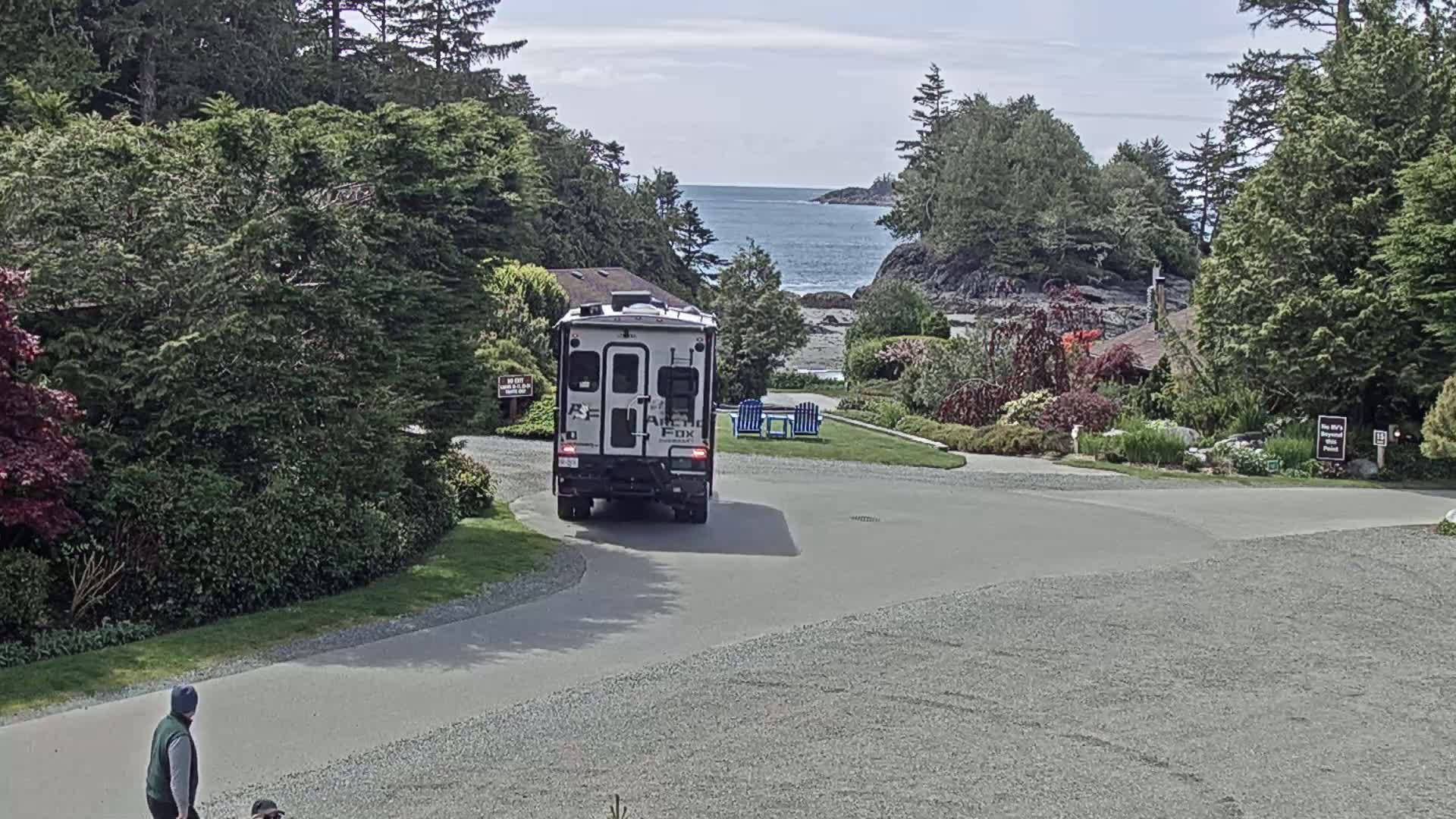 A motorhome backs out of a parking area onto a gravel road, with a person walking in the foreground, overlooking a calm ocean under a partly cloudy sky.