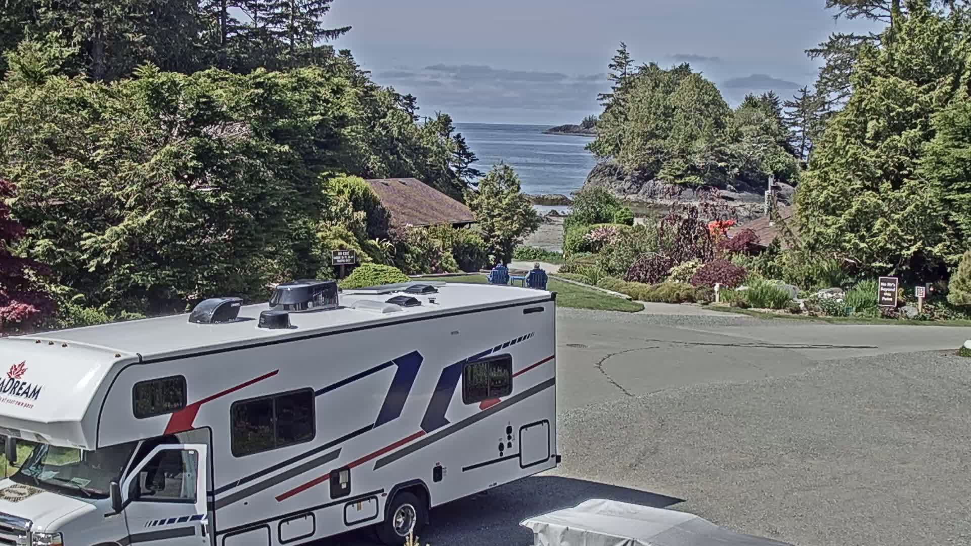 A white camper van is parked in a paved area overlooking a calm ocean bay, surrounded by lush green trees and landscaping on a sunny day.