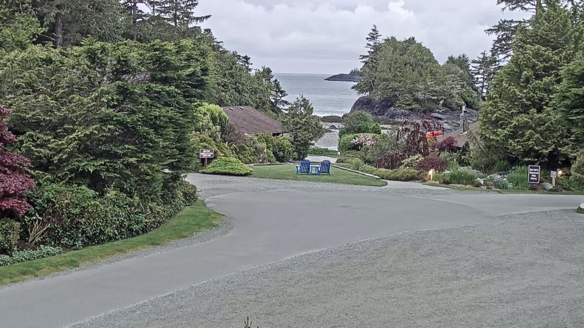 A paved road curves through a landscaped area with green lawns, shrubs, and trees, leading to a view of a calm ocean under a cloudy sky.