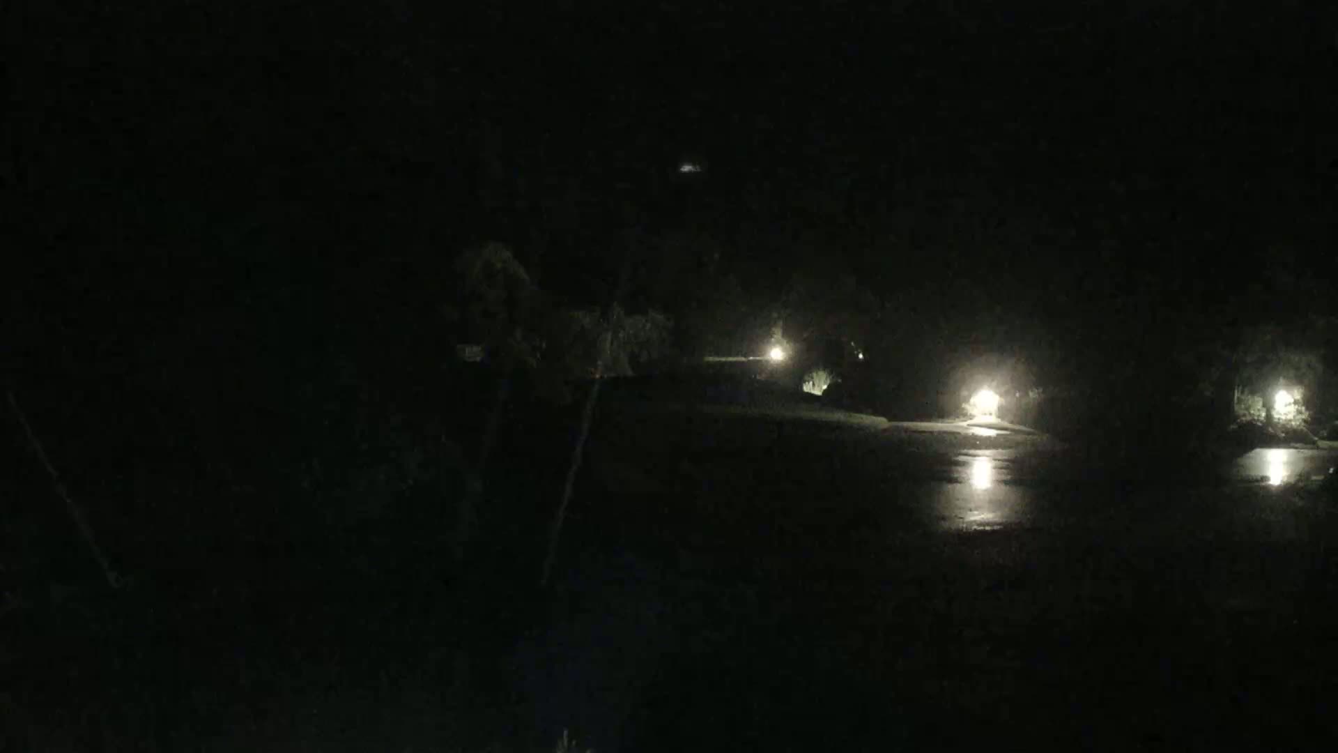 A dark, nighttime scene shows a body of water reflecting the lights of distant buildings and trees under a clear sky.