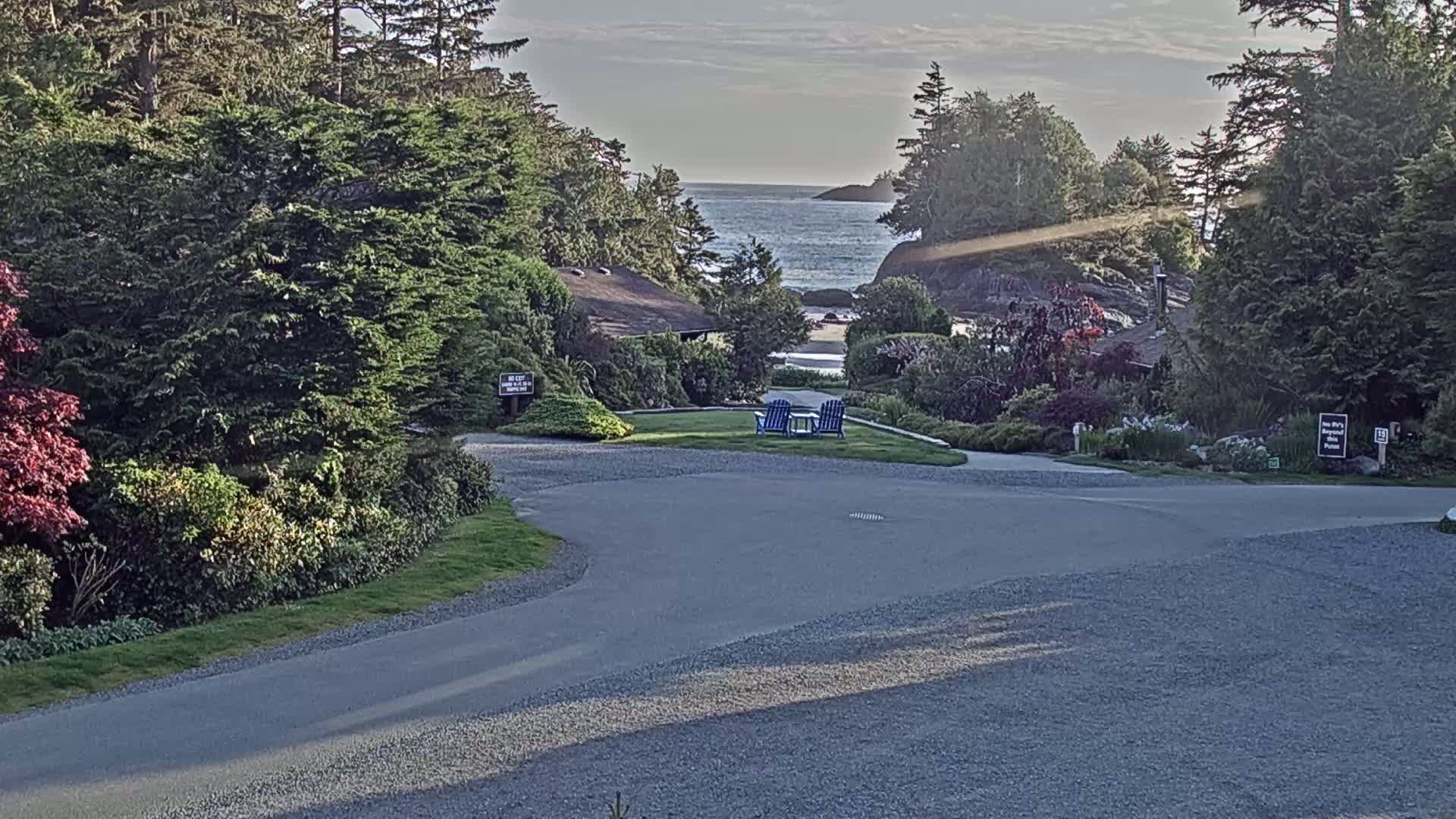 A paved driveway curves through a landscaped area with lush green vegetation, leading to a view of a calm ocean under a partly cloudy sky; two blue chairs sit on a small lawn in the midground.