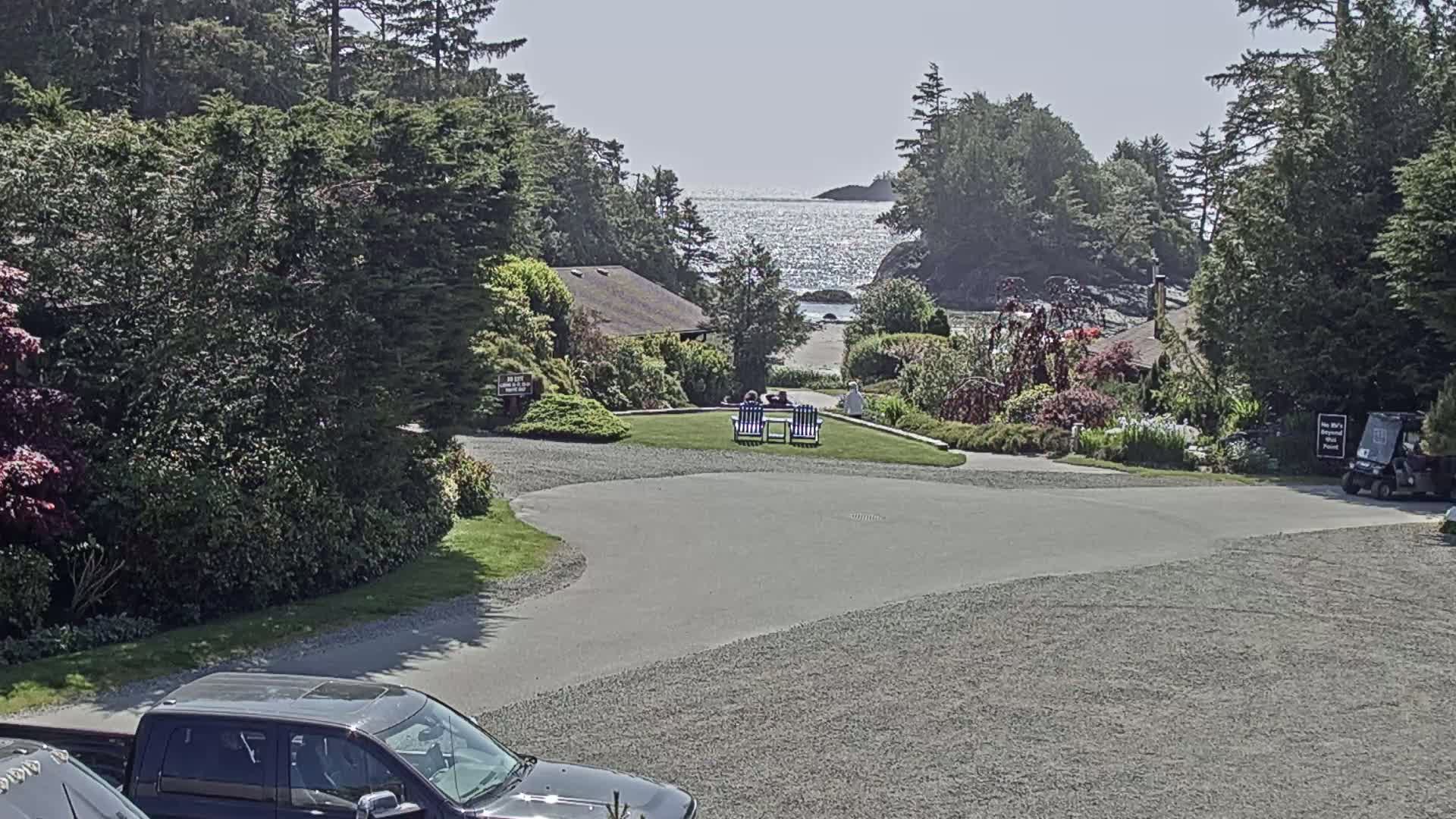 A gravel parking lot overlooks a grassy area with two people sitting in chairs, a small building, lush green vegetation, and a sparkling ocean under a sunny sky.
