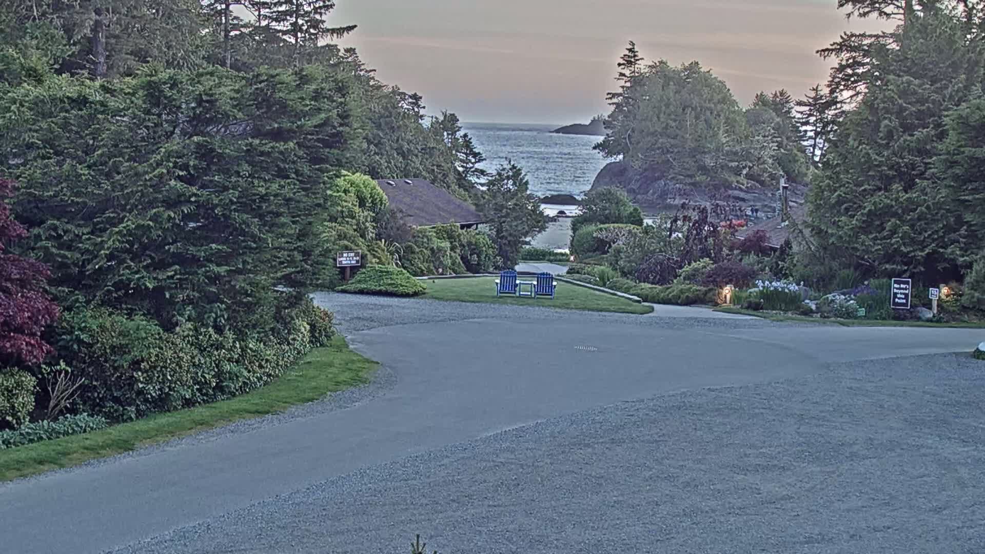 A paved driveway curves through a lush, green landscape toward a calm ocean view under an overcast sky, with two blue chairs positioned on a grassy area in the mid-ground.