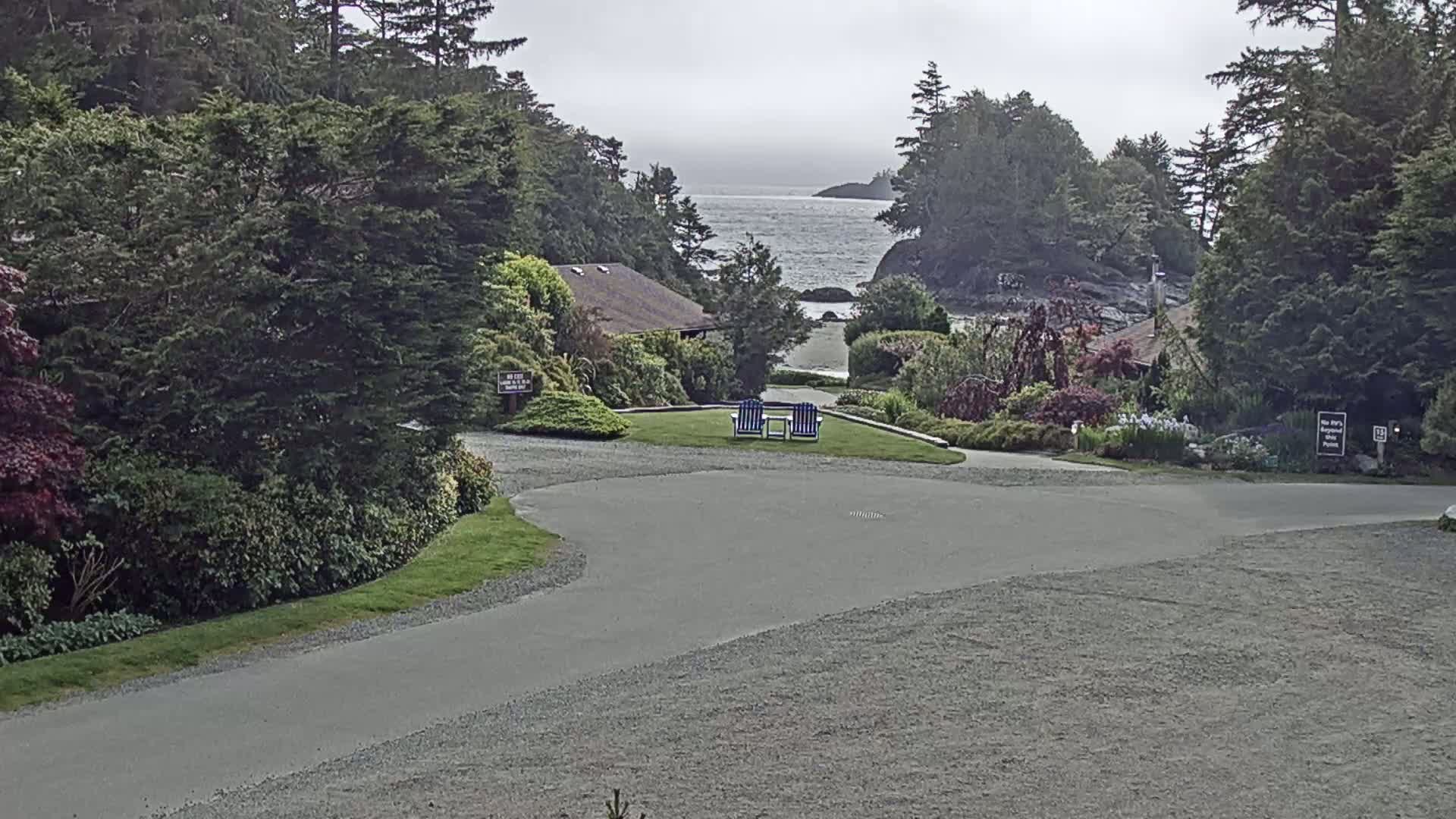 A paved driveway curves through a lush, green landscape toward a calm ocean visible in the distance under an overcast sky, with two blue chairs situated on a grassy area nearby.