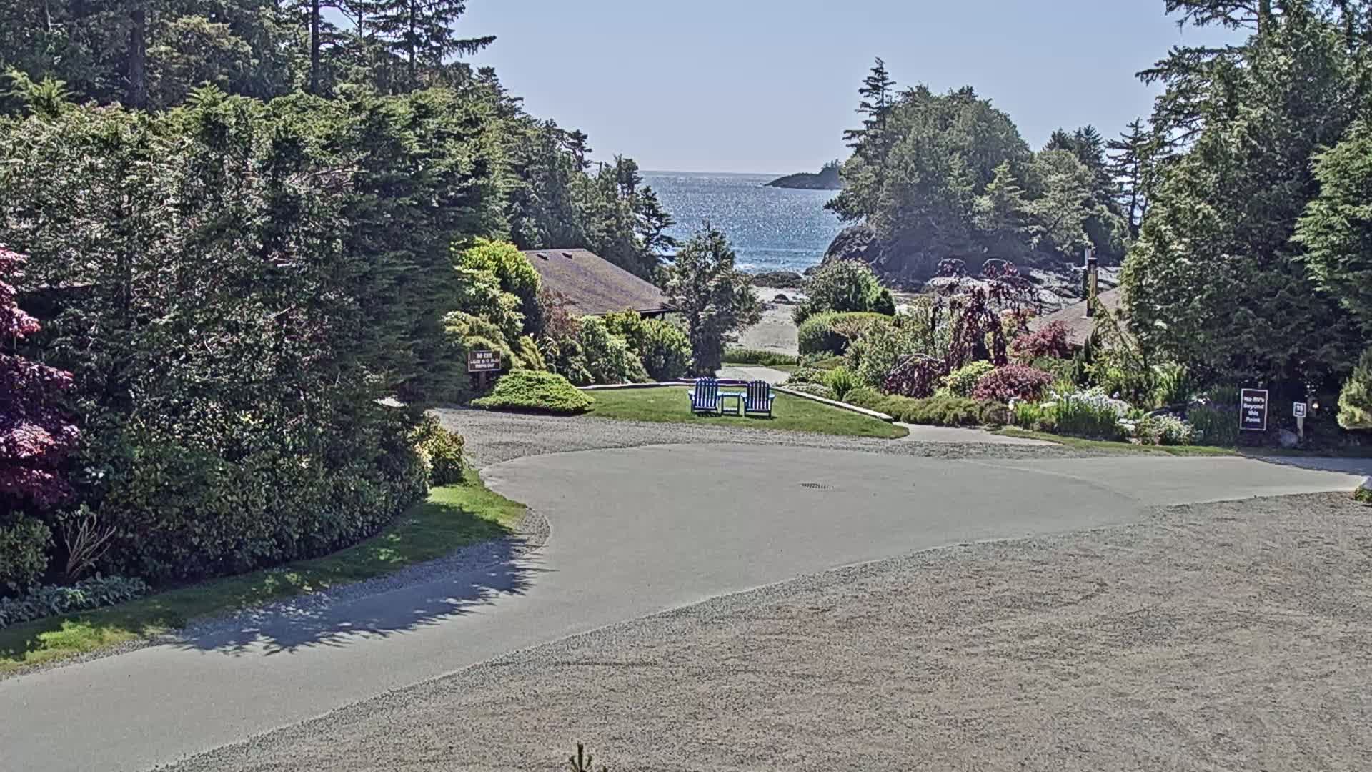 A paved roadway curves through a landscaped area with buildings, lush greenery, and a view of a calm ocean beyond a sandy beach under a sunny sky.