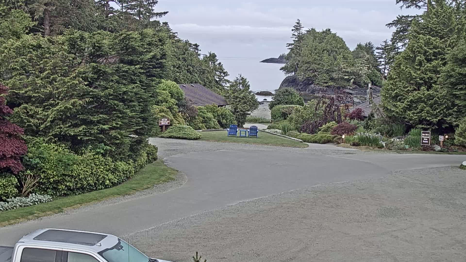 A paved driveway curves through a lush, green landscape with a view of a calm ocean beyond, under a mostly cloudy sky.