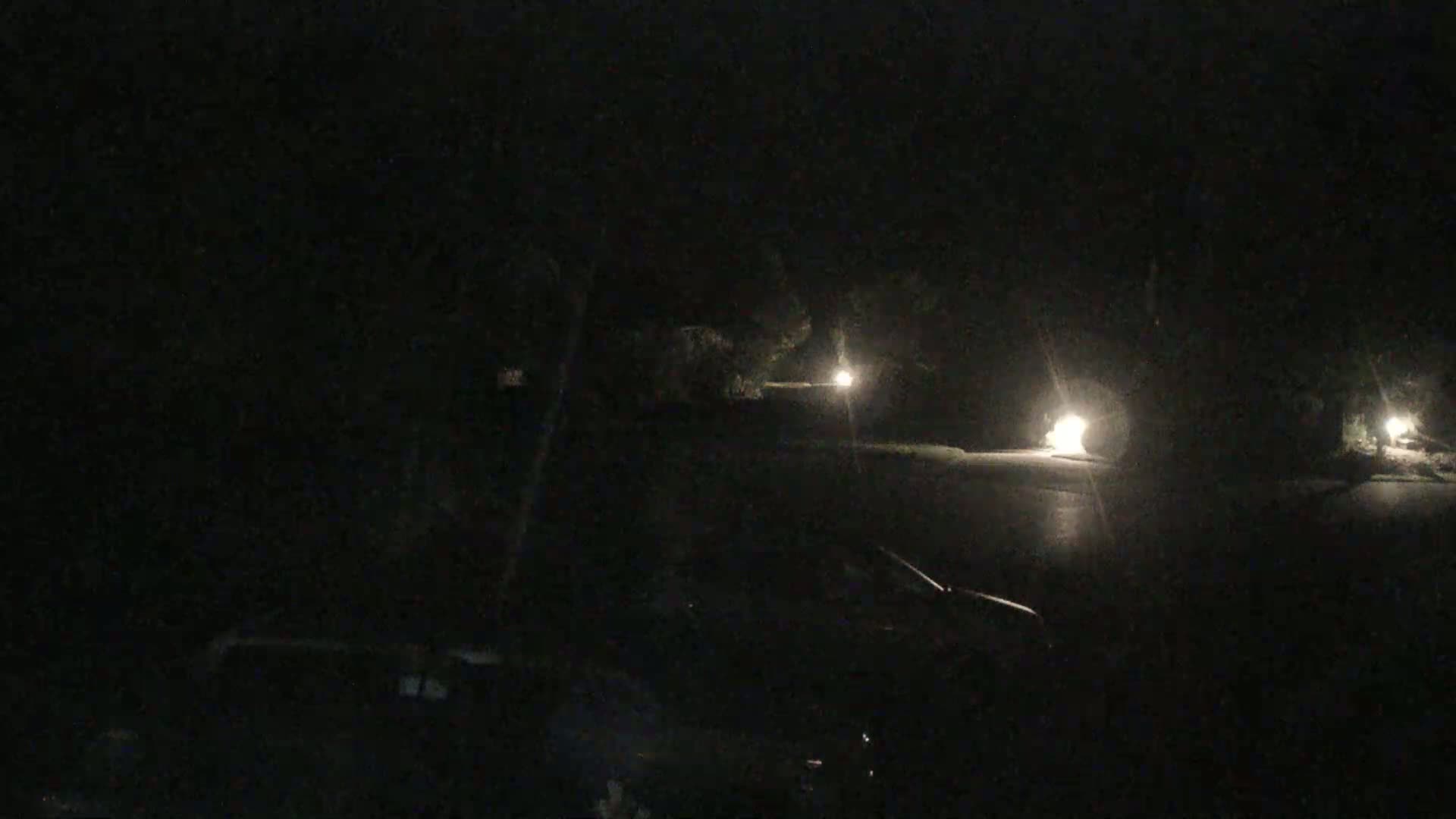 A dark, nighttime scene shows a street with dimly lit houses and a vehicle in the foreground.