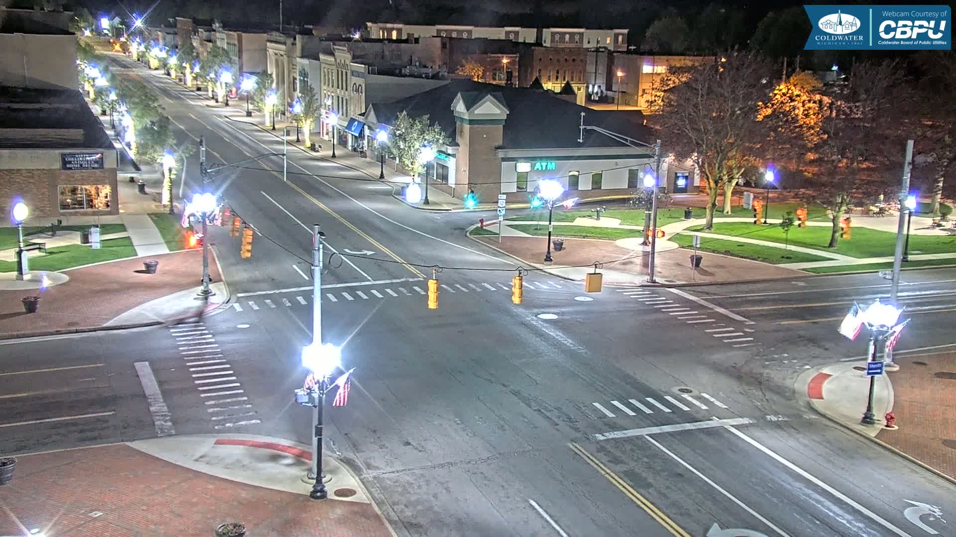 A nighttime aerial view of a town square intersection with illuminated streets, buildings, and landscaping.