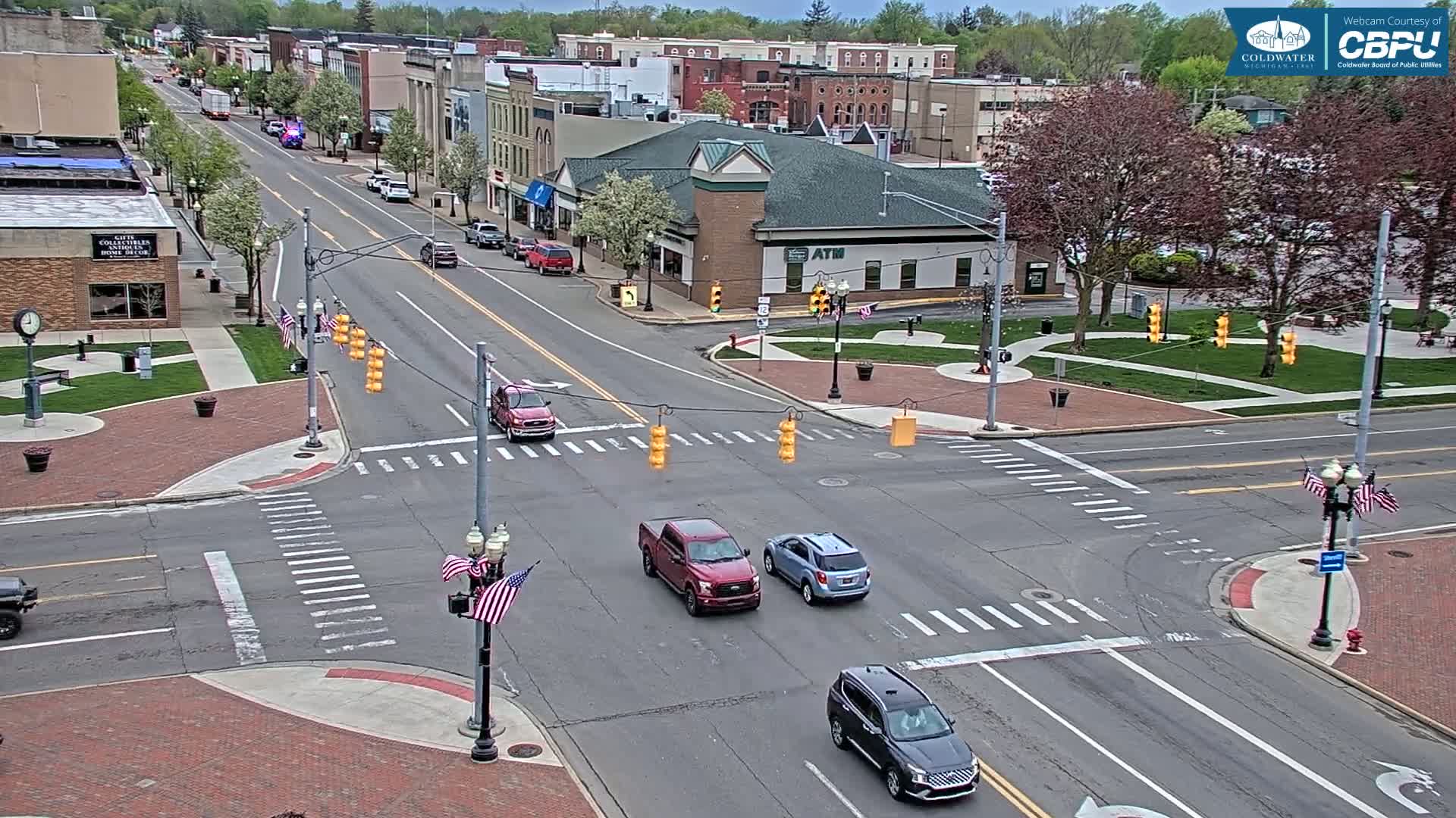 A partly cloudy day reveals a town square intersection with several vehicles at a four-way stop, surrounded by brick sidewalks, green lawns, and buildings.
