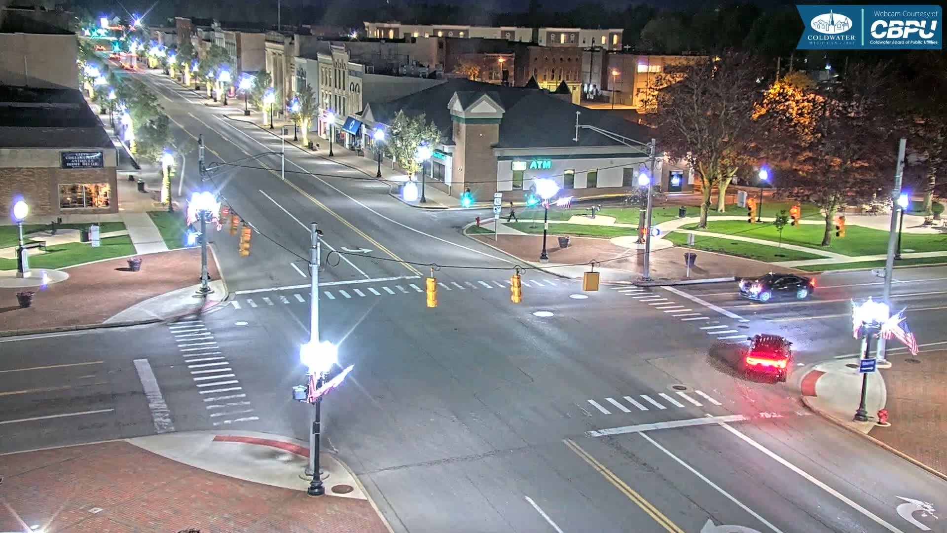 A nighttime aerial view shows a lightly trafficked street intersection in a town with buildings lining the street, streetlights illuminating the scene, and a small park visible in the background.
