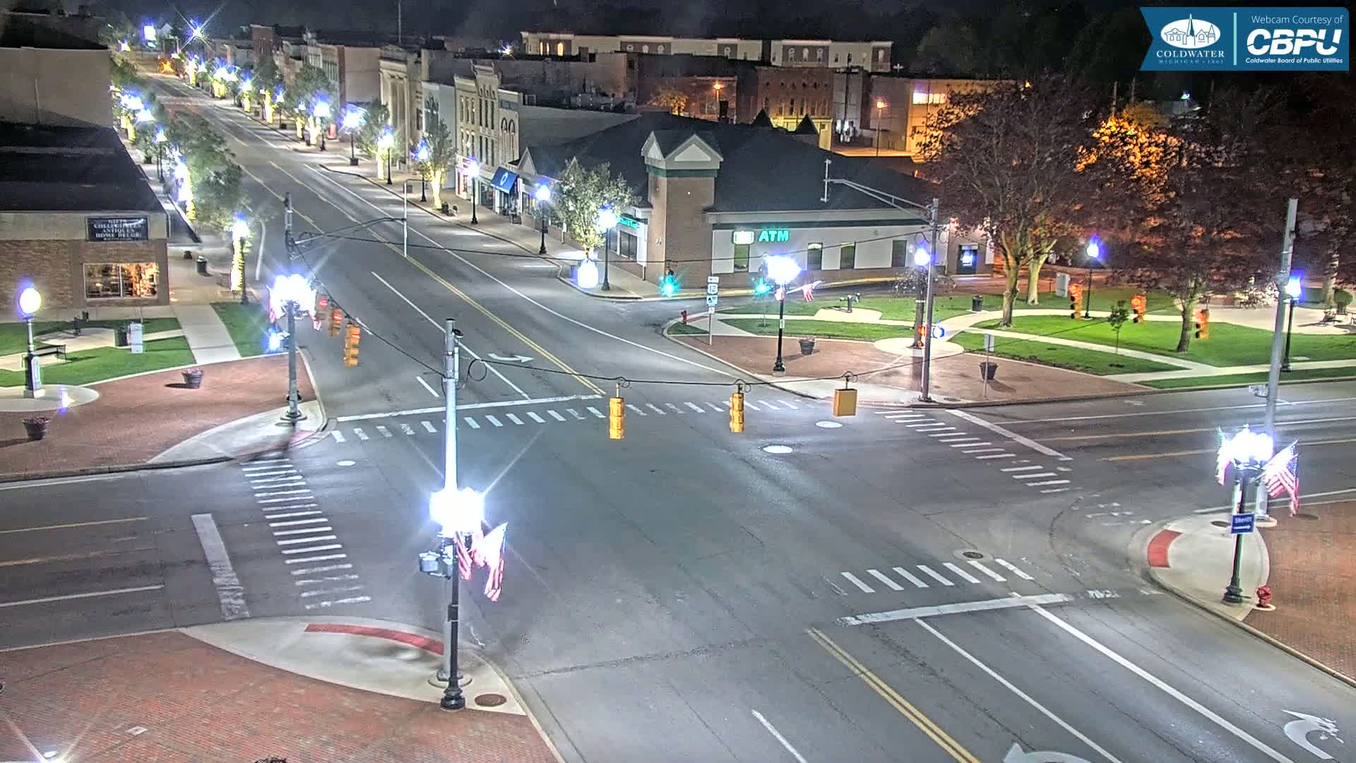 A nighttime, illuminated view of a town square intersection with several buildings lining a street, featuring crosswalks, traffic lights, and trees in the surrounding area.
