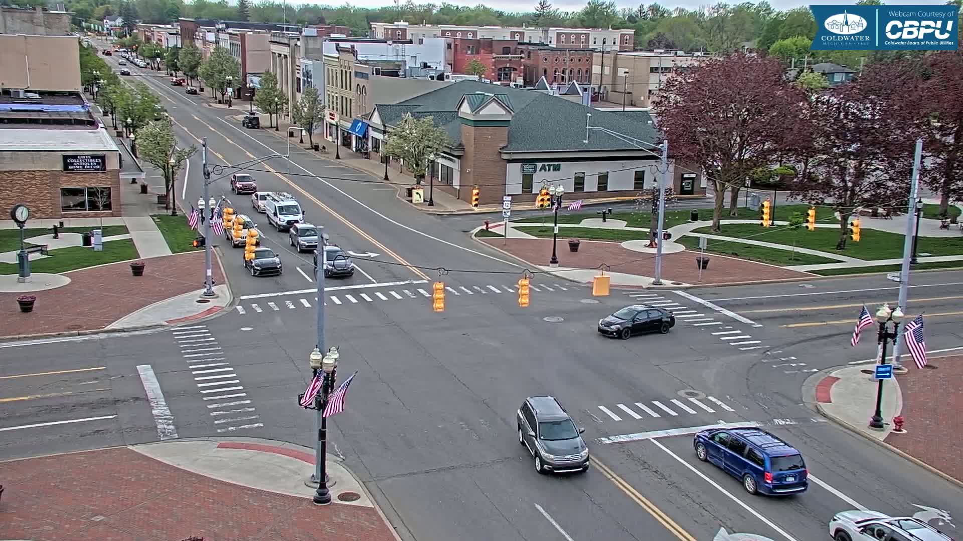 A street intersection in a town on a partly sunny day, with several cars at a traffic signal and some parked along the sides of the road, and American flags displayed near the street corners.