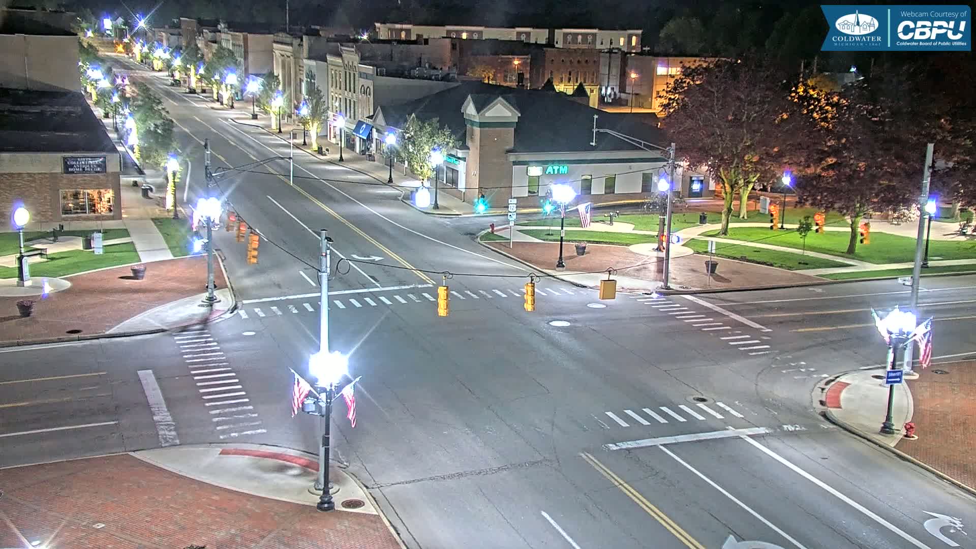 A nighttime aerial view of a town square intersection with brick sidewalks, streetlights, and trees, showing relatively little traffic.