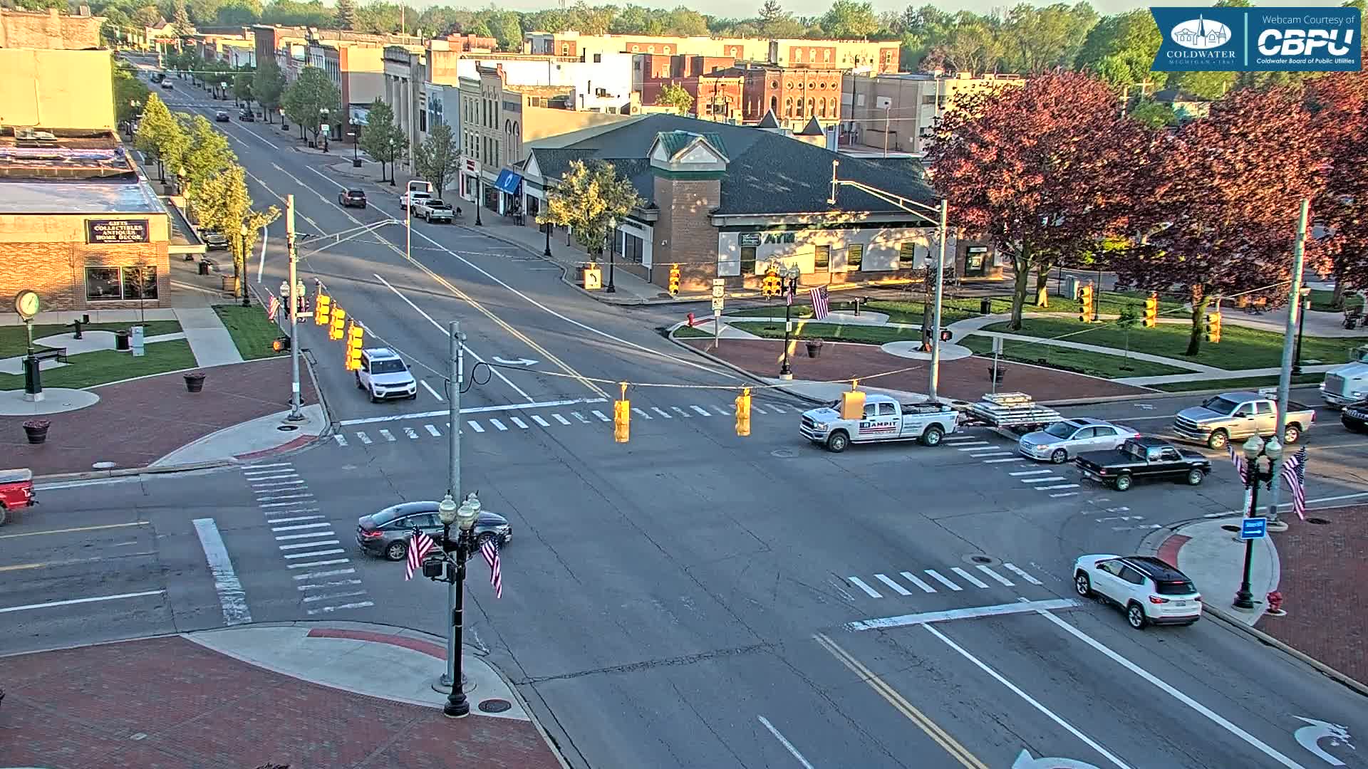 A sunny, daytime view of a town square intersection with several vehicles at a four-way stop, surrounded by brick sidewalks, trees, and buildings.