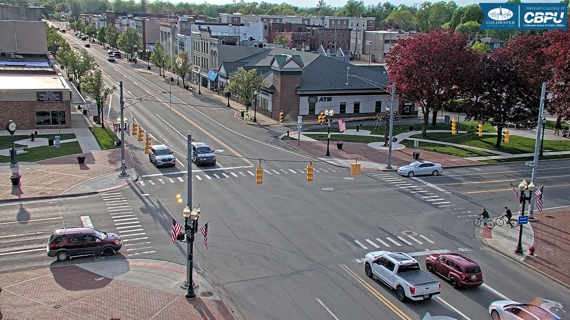A sunny day view of a town's street intersection with several vehicles and two cyclists crossing, surrounded by buildings and lush green trees.