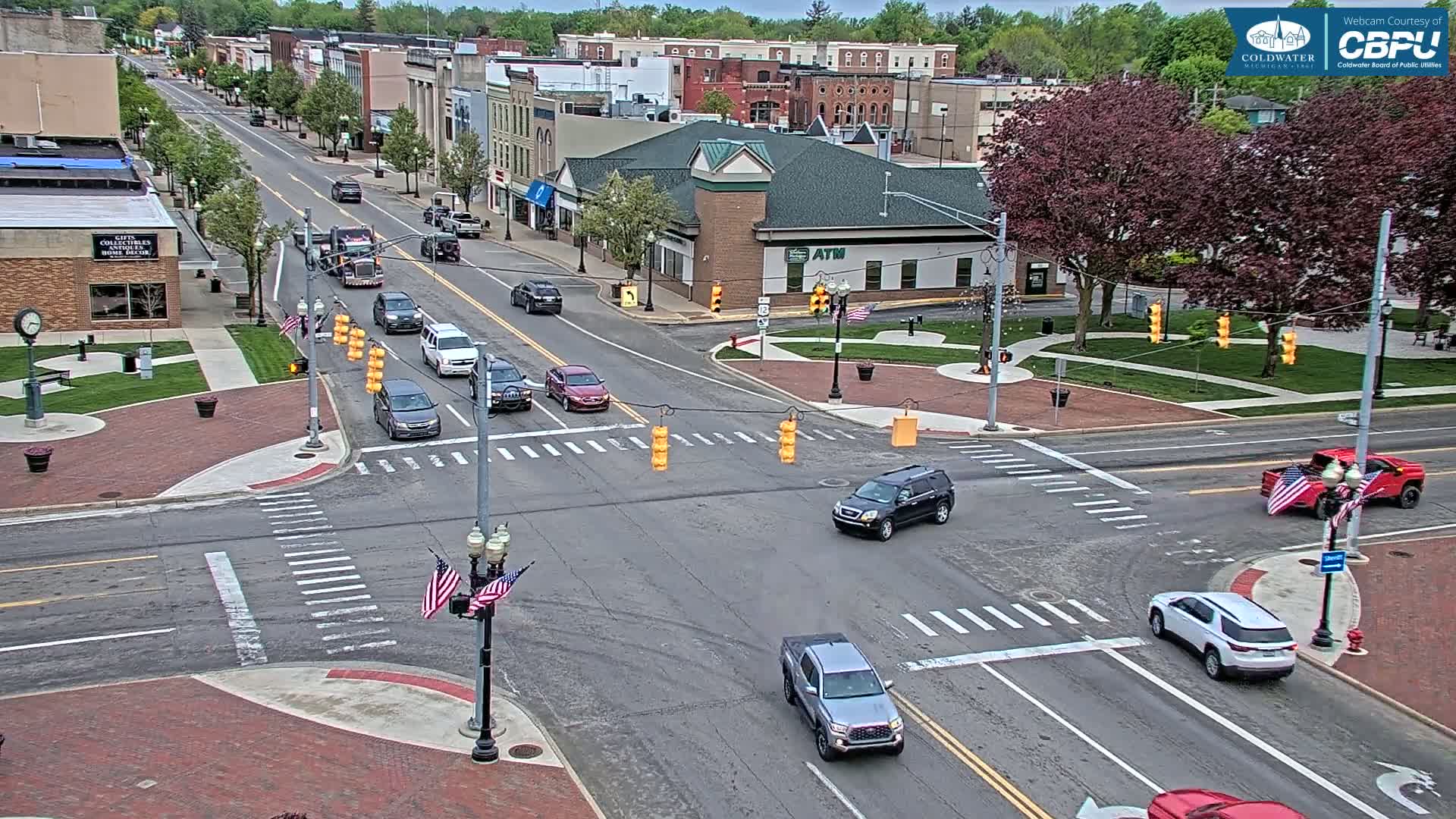 A mostly sunny overhead view of a town intersection with several cars at a traffic light, surrounded by brick sidewalks, buildings, and lush green trees.