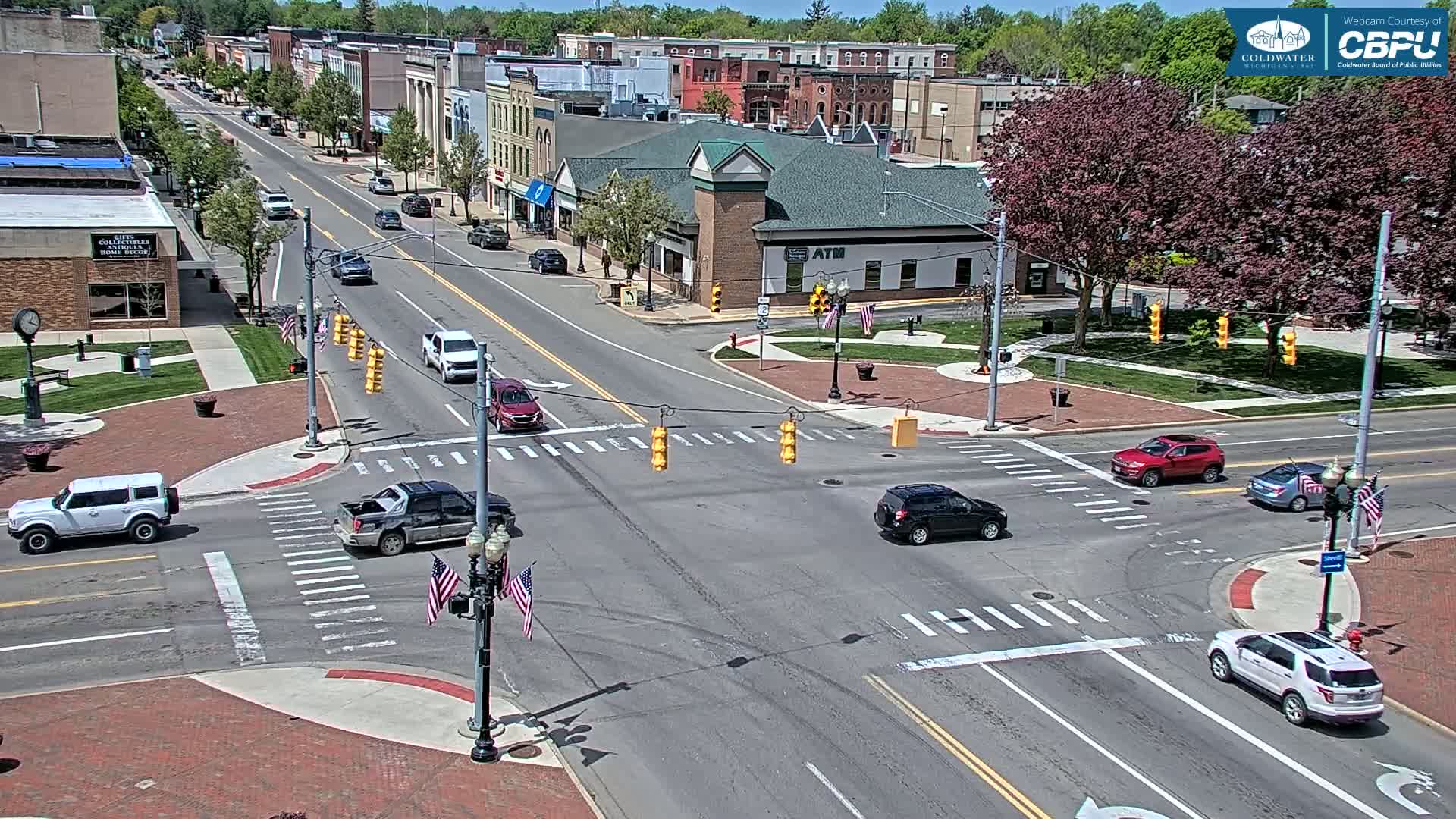 A sunny day reveals a street intersection in a town with several cars at a traffic light, surrounded by brick buildings and lush green trees.
