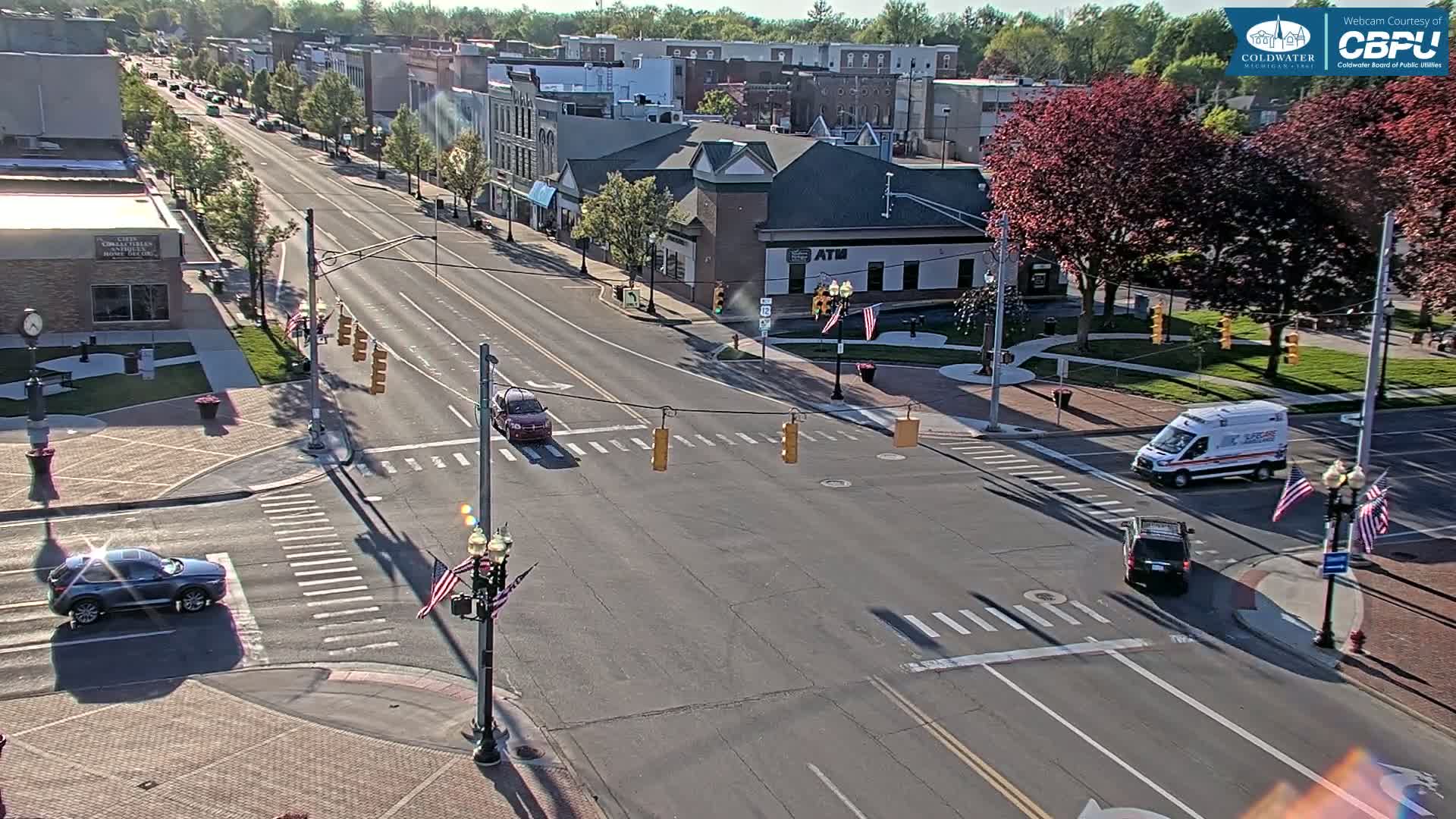 A sunny day shows a mostly empty four-lane road intersecting with a cross street in a town square, with a few cars and an ambulance visible.