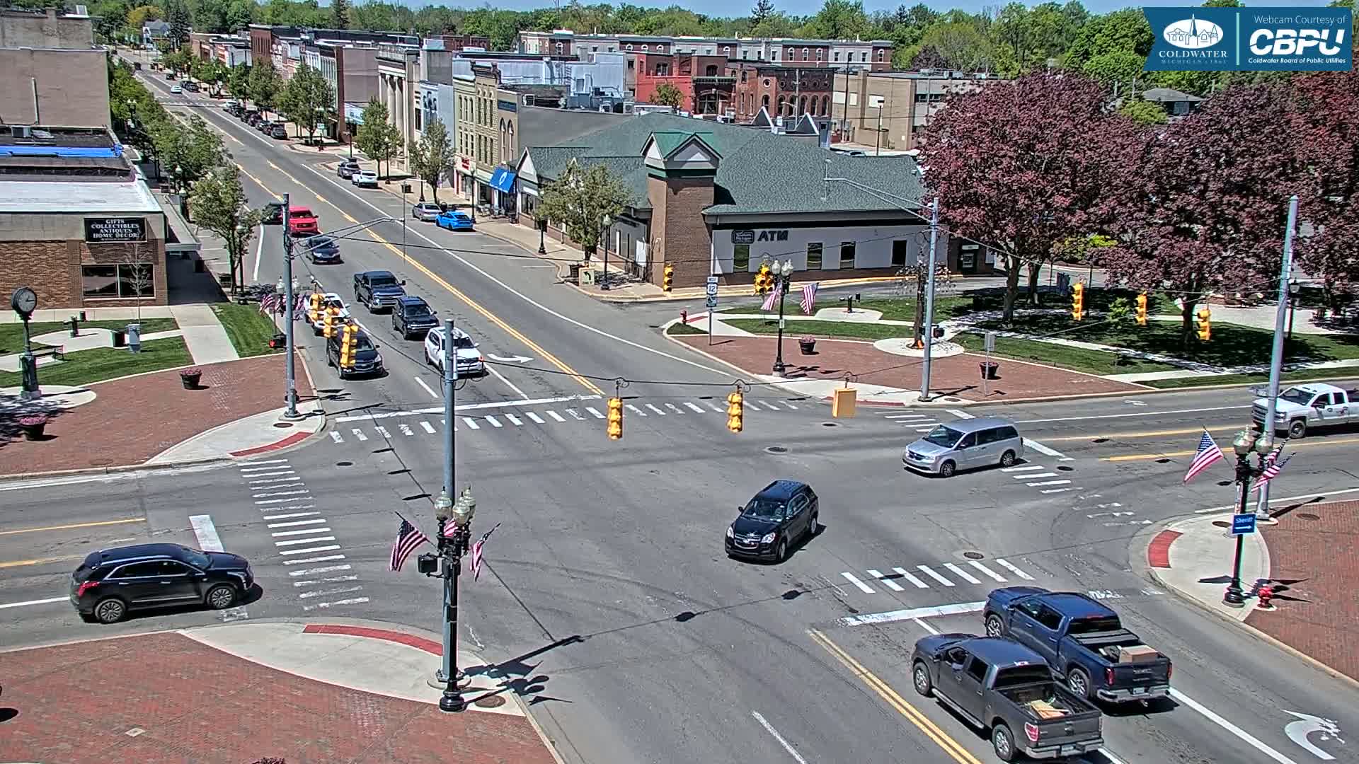 A sunny day shows a town square intersection with several vehicles, including cars and pickup trucks, at a four-way stop with traffic signals; multiple buildings and trees surround the intersection.