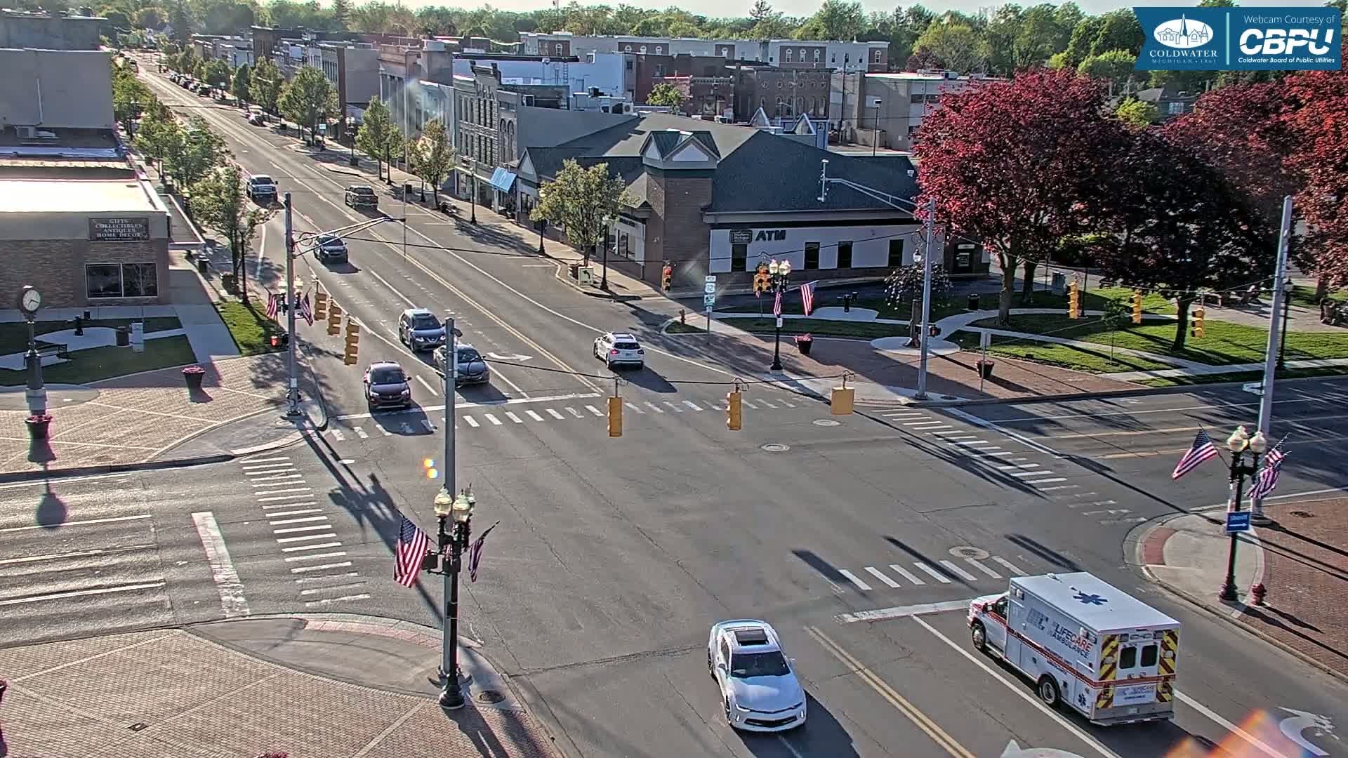 A sunny day reveals a street intersection in a town with several cars, an ambulance, and American flags on light posts, with buildings and trees lining the streets.