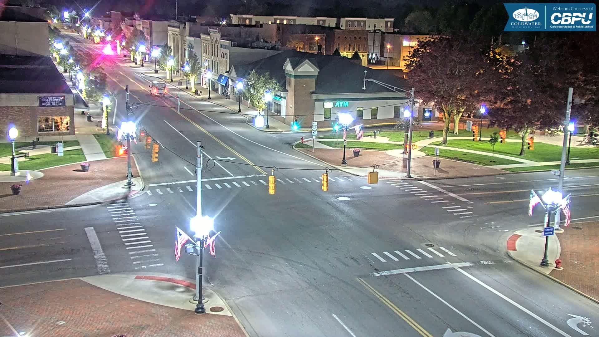 A nighttime, high-angle view of a town square intersection with several cars on the road, streetlights, and small American flags displayed on light posts.