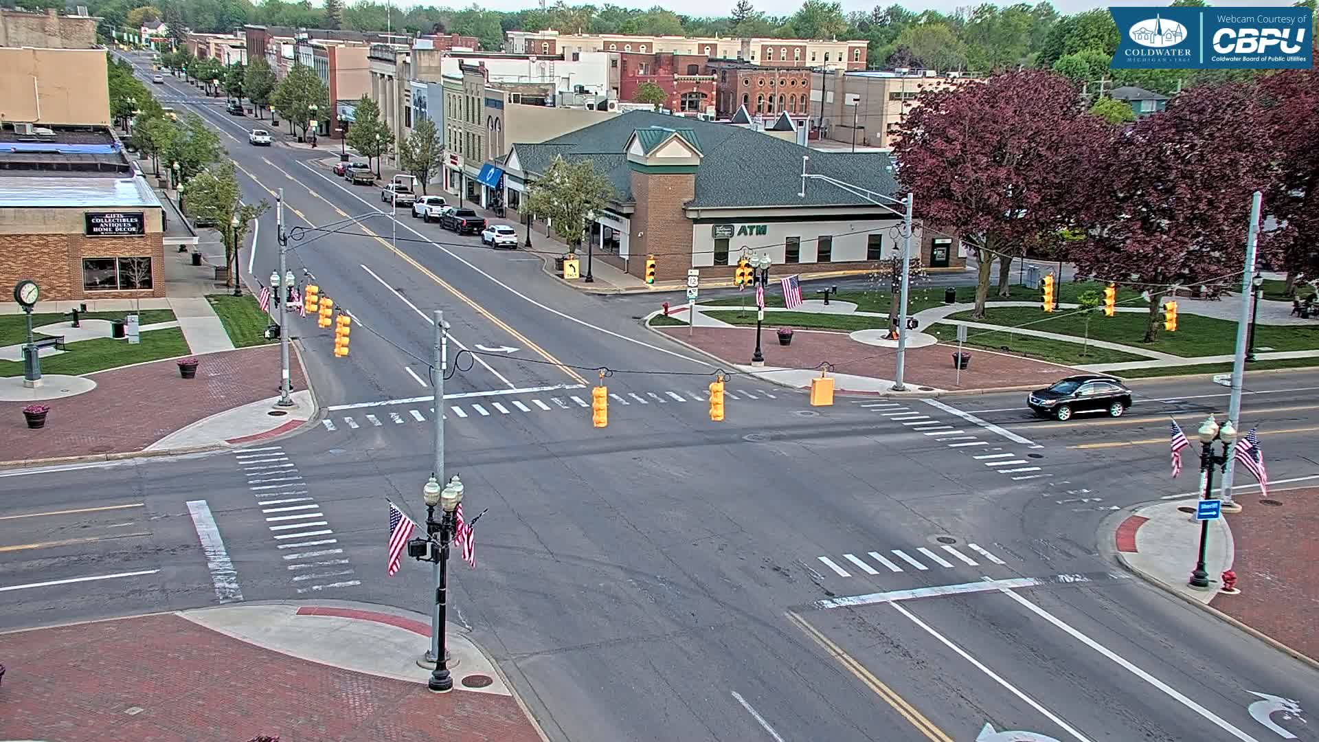 A mostly sunny day shows a street intersection in a town with brick sidewalks, buildings, and lush green trees; a single dark-colored SUV is visible on the road.