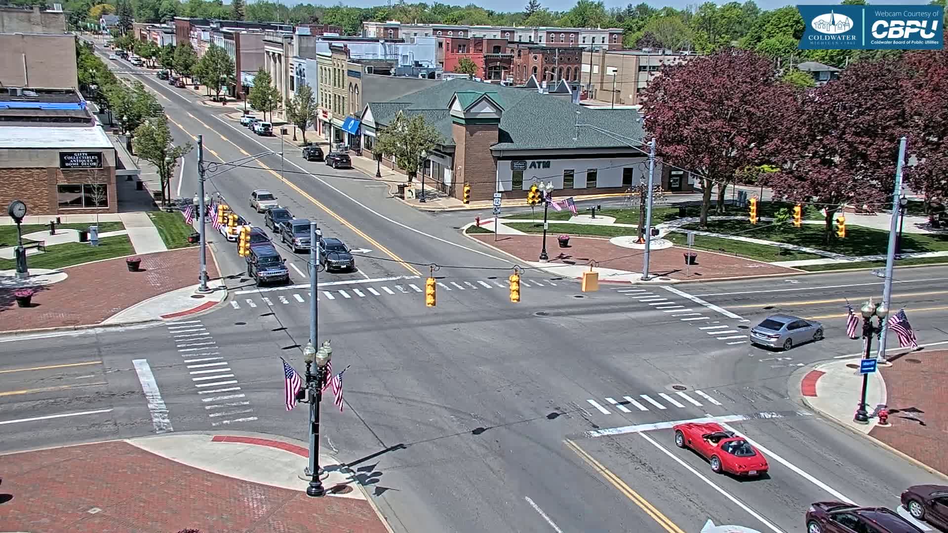 A sunny day reveals a town square intersection with several cars and small American flags displayed on light posts.