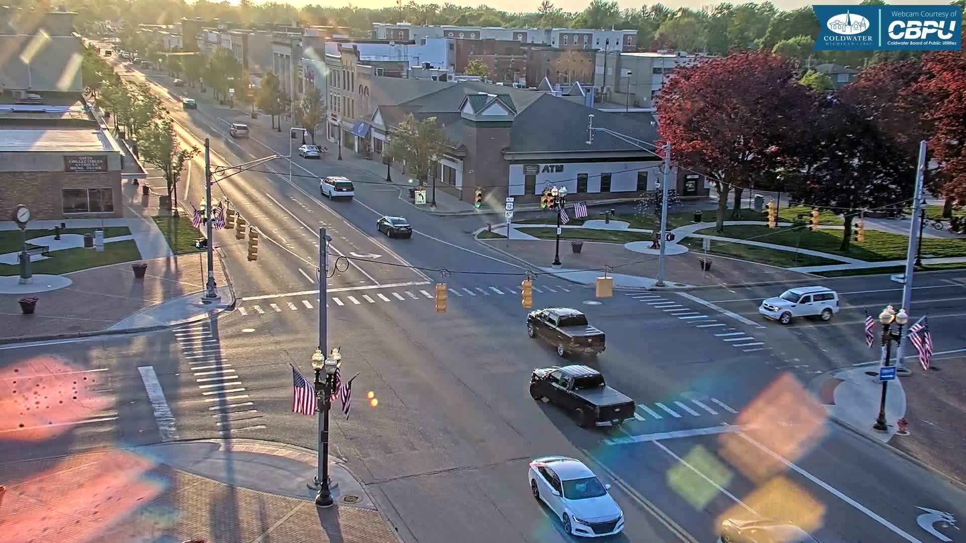 A sunny, mostly clear day reveals a high-angle view of a town's intersection with several cars,  American flags, and buildings lining a main street.